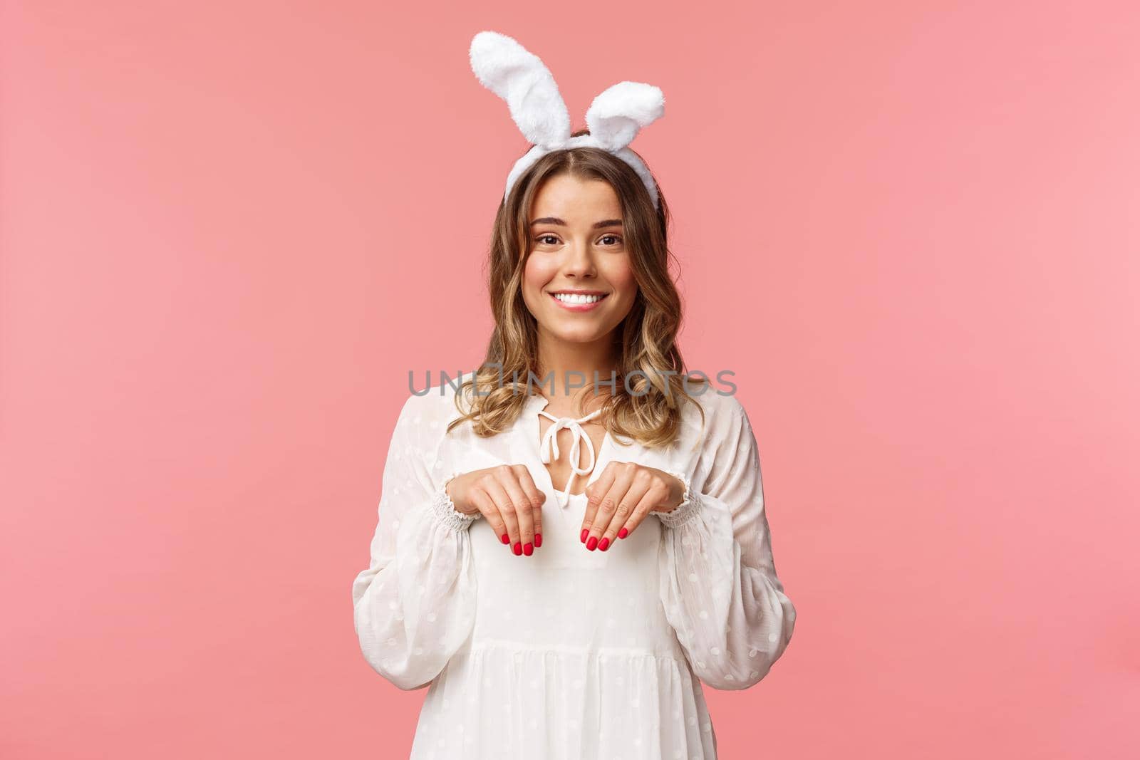 Holidays, spring and party concept. Portrait of cute and tender, lovely blond girl imitating bunny, holding hands like paws and wearing rabbit ears, smiling camera, pink background.