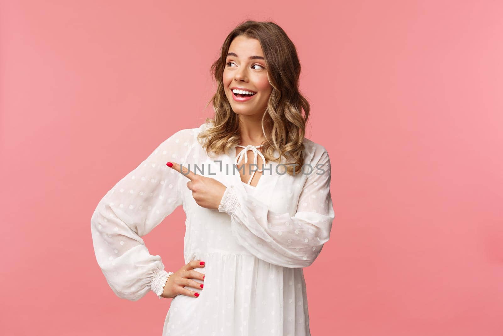 Curious upbeat beautiful blond woman enjoying summer feeling cheerful, pointing looking left with interested, enthusiastic expression, wear cute white dress, standing pink background.