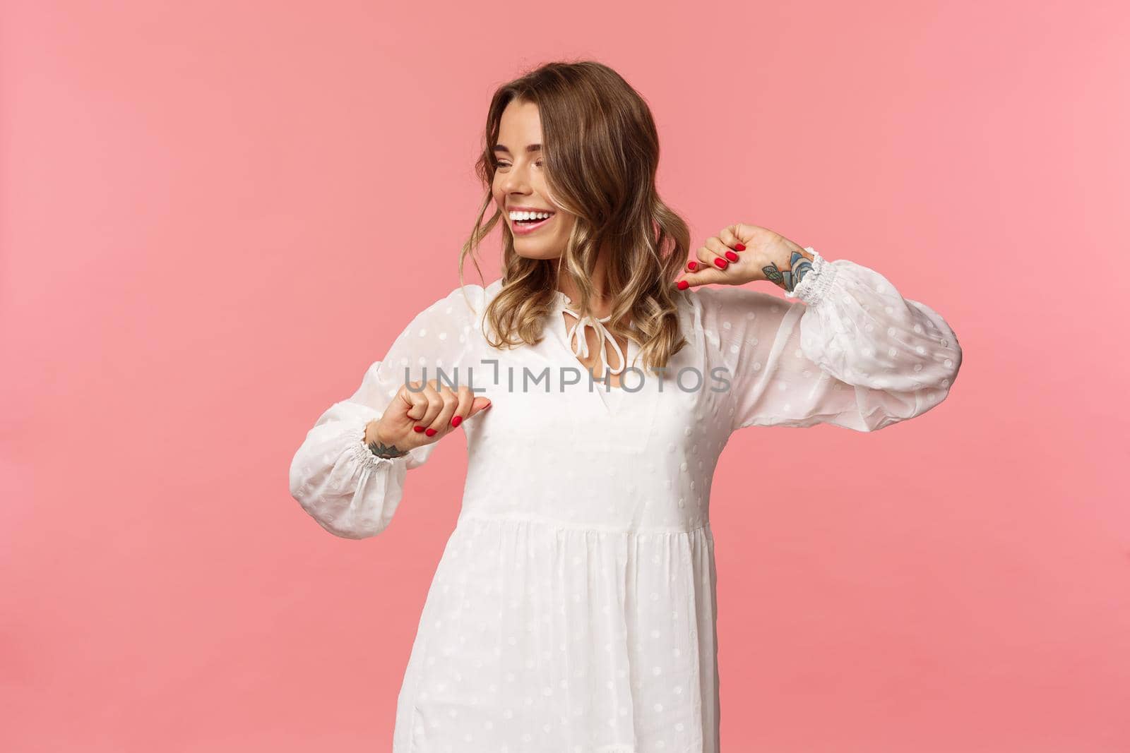 Beauty, fashion and women concept. Tender and carefree pretty young girl enjoying spring time, wearing white dress, dancing and looking away with beaming smile having fun, pink background.