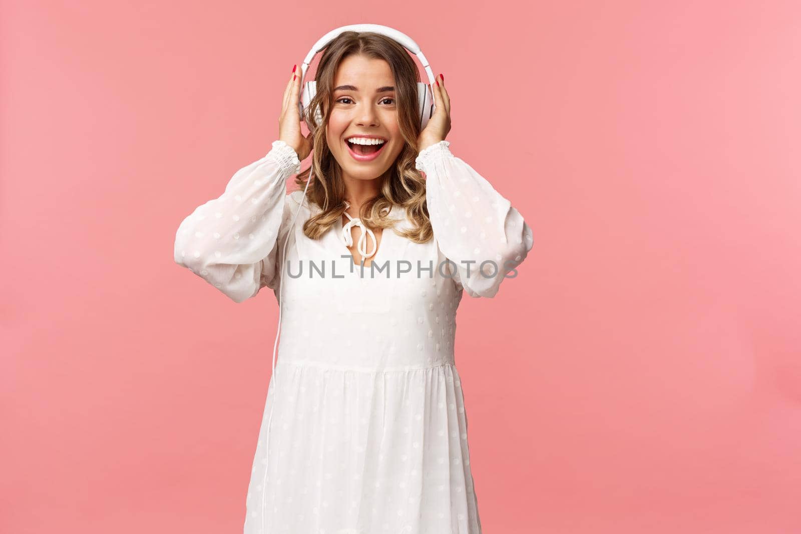 Portrait of excited, happy good-looking girl in white tender dress, wearing headphones and smiling amazed as looking at camera, fascinated with good sound quality, pink background by Benzoix