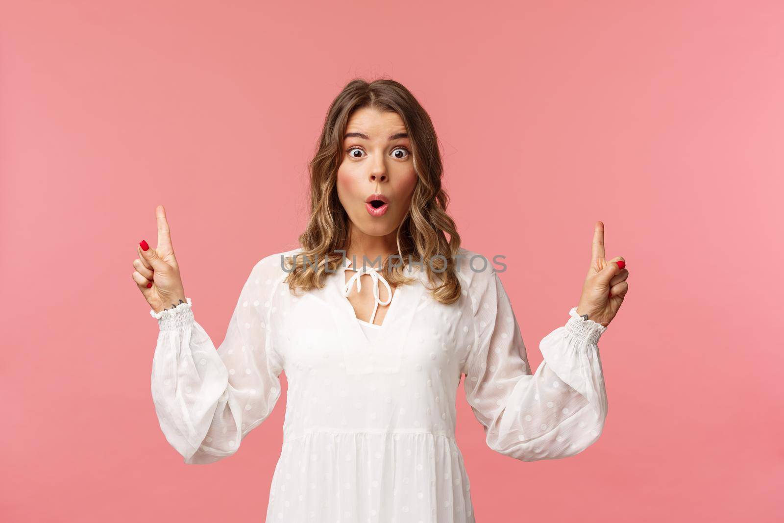 Waist-up portrait of amazed blond girl say wow in excitement, pointing fingers up, showing person top advertisement with cool special spring discount, standing pink background by Benzoix