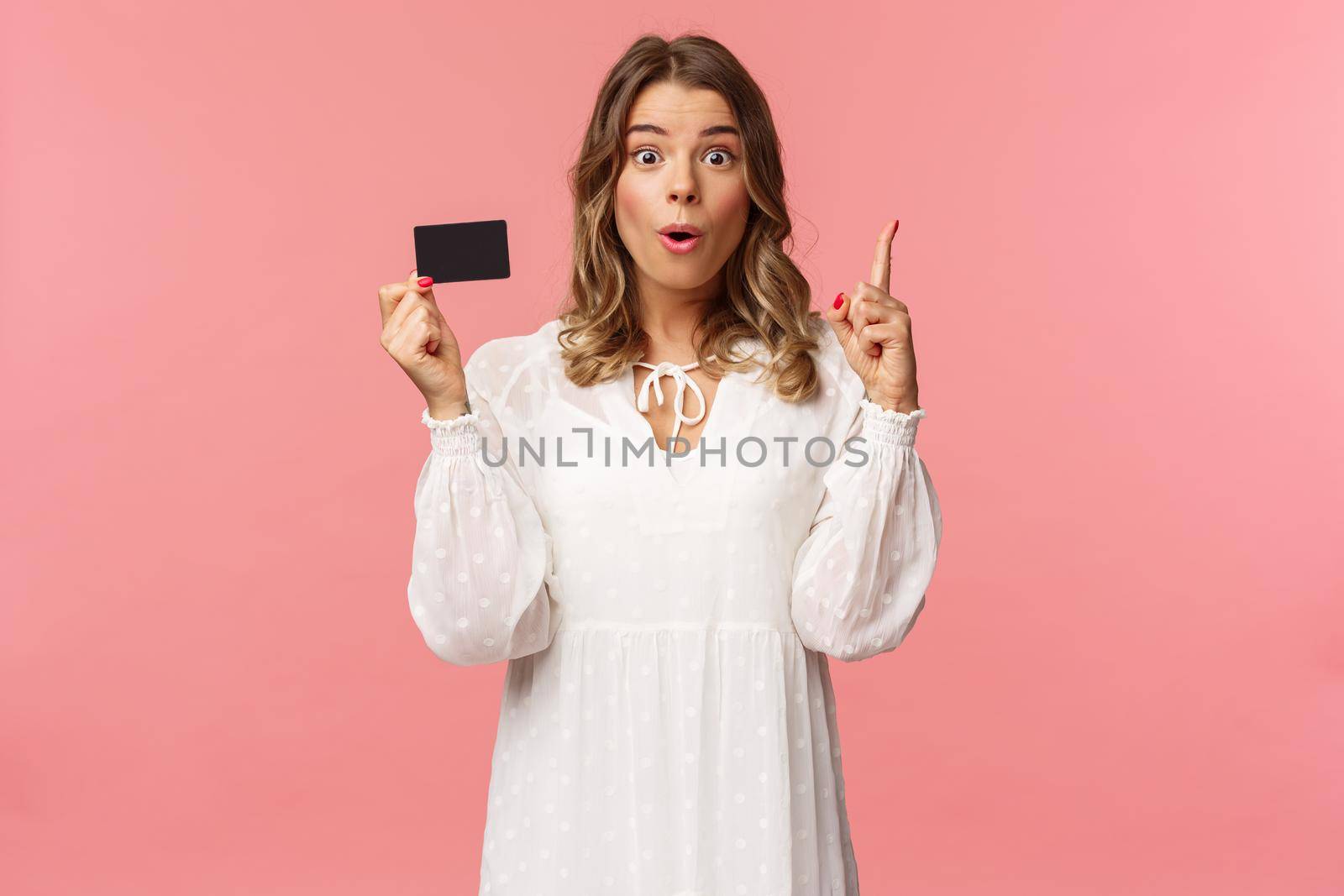 Portrait of thoughtful cute blond girl in white dress, have and excellent idea, holding credit card say wow, eureka, raise index finger have suggestion, know where find needed product by Benzoix