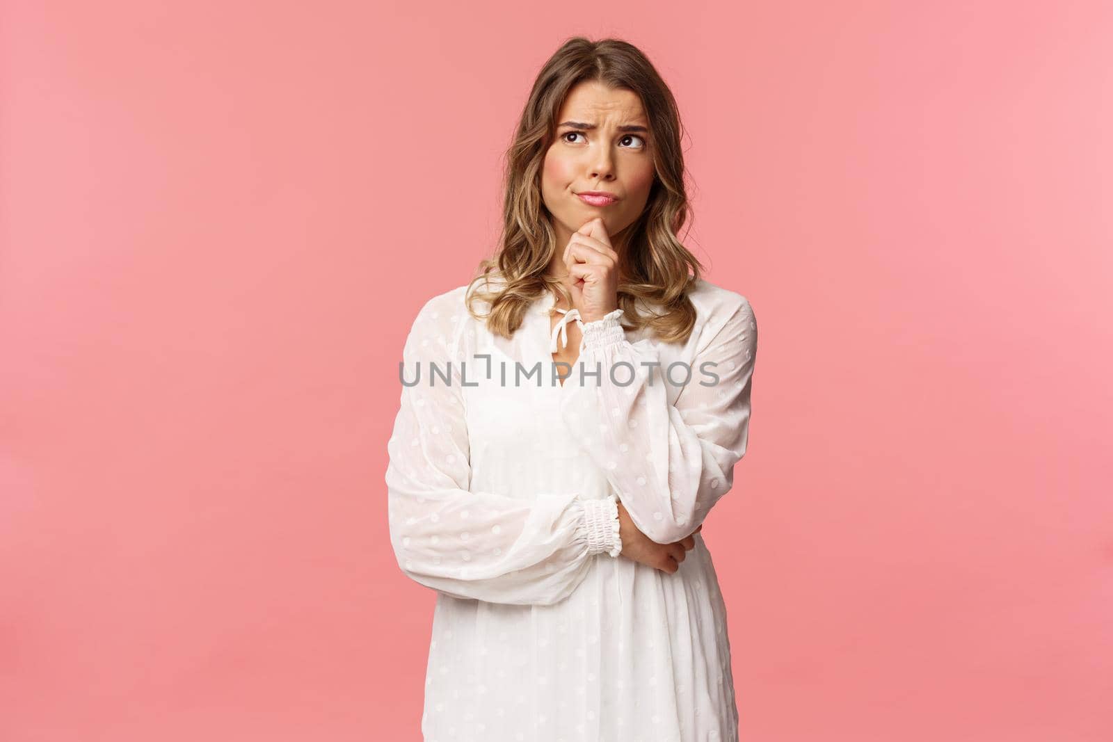 Beauty, fashion and women concept. Portrait of uncertain, serious-looking feminine young blond woman in white dress, touch chin, smirk and frowning thoughtful, look away thinking.