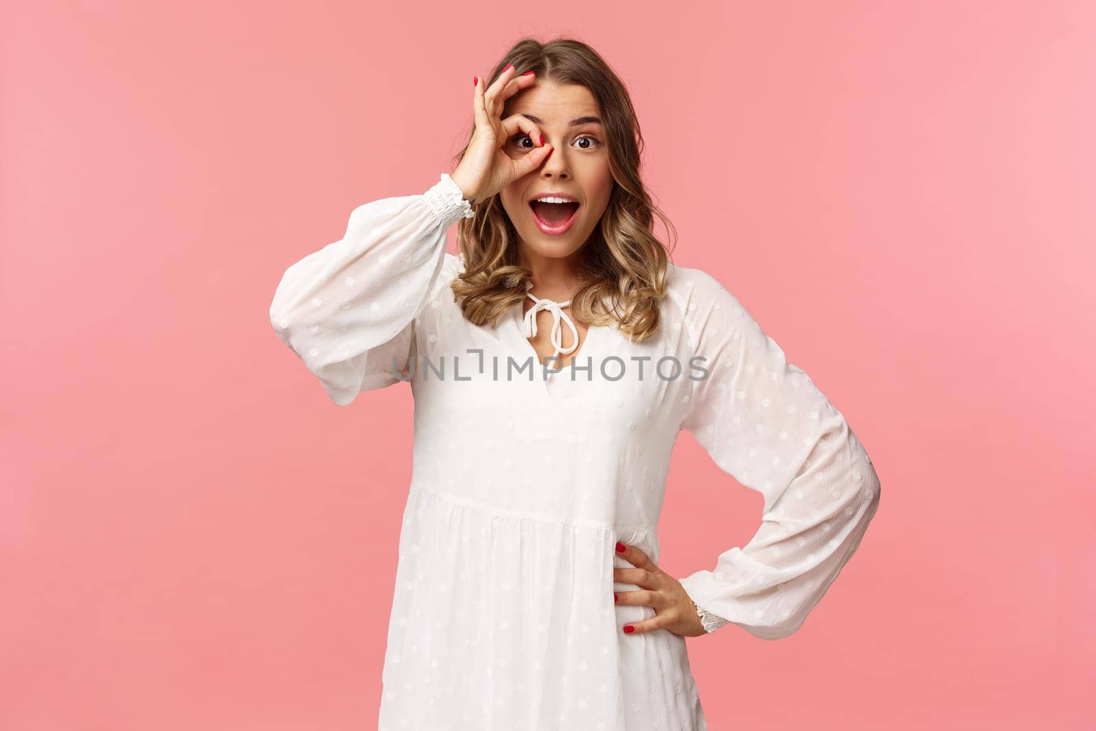 Portrait of amazed and fascinated, surprised young blond girl seeing something really cool, stare with excited and happy face, smiling found what looking for, look through okay sign over eye.