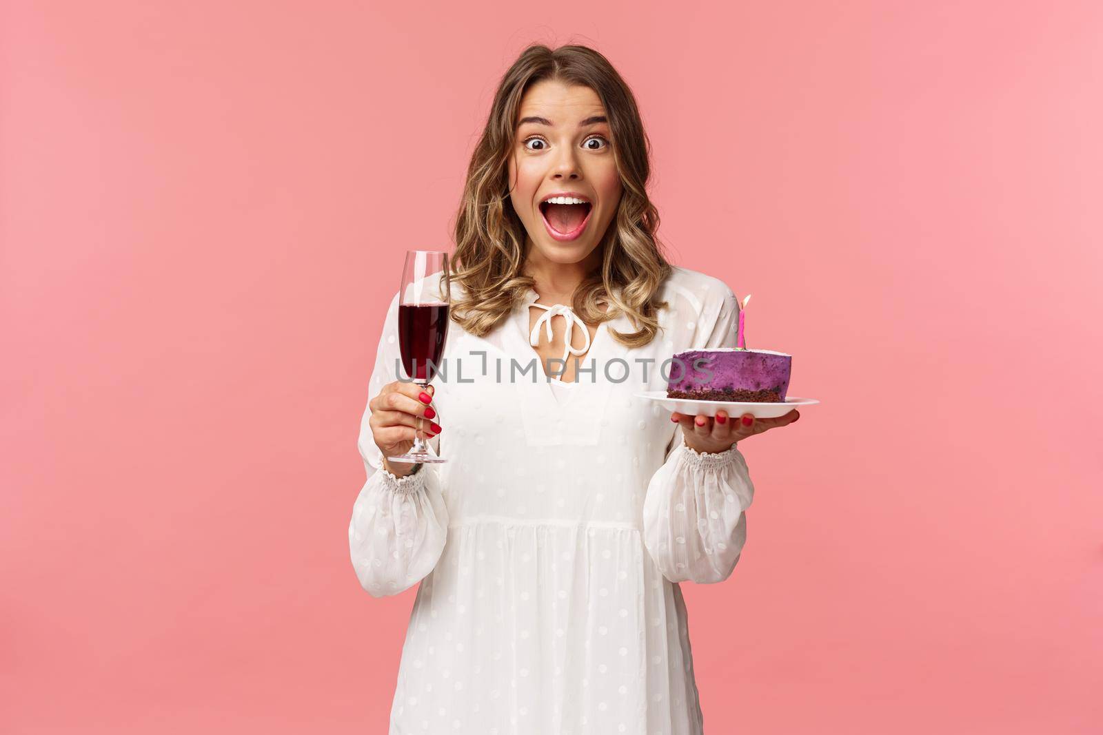 Holidays, spring and party concept. Portrait of fascinated and amused blond girl in white dress look astonished camera, celebrating birthday with b-day cake and glass of wine, pink background.