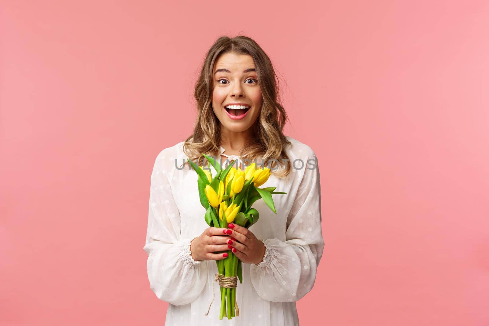 Holidays, beauty and spring concept. Portrait of excited and amazed young beautiful blond girl in white dress, holding yellow tulips, receive flowers as gift, smling amused, pink background by Benzoix