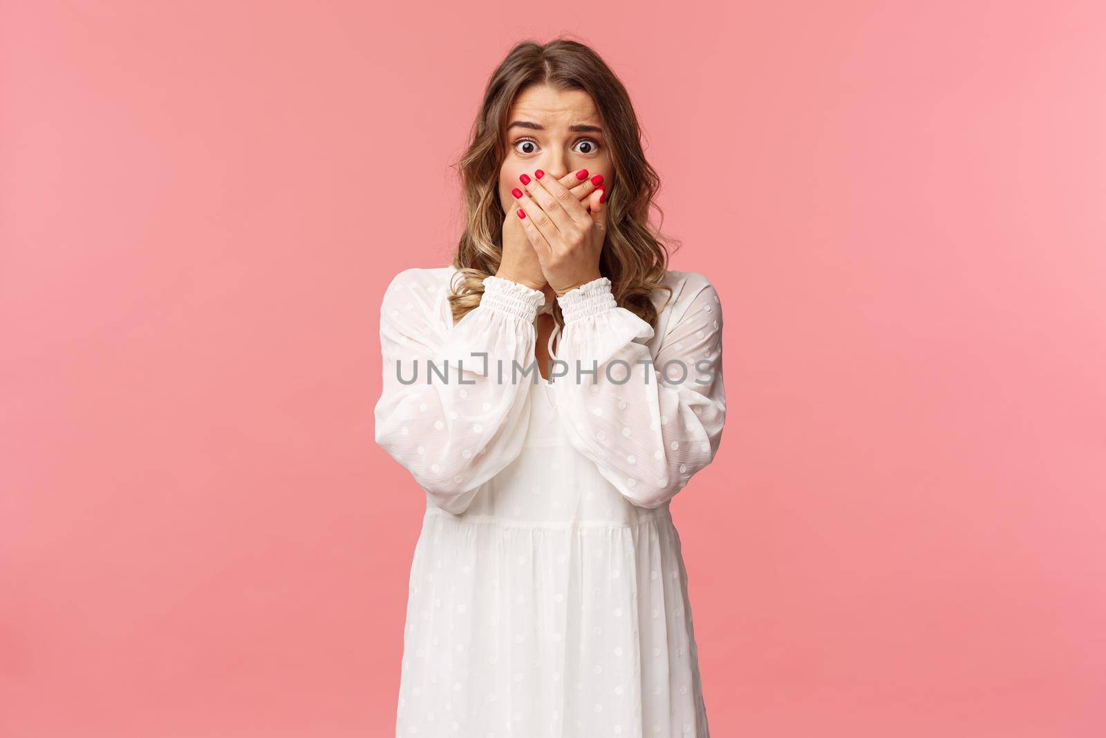 Shocked and anxious young concerned pretty girl in white dress, gasping close mouth with hands, look worried and startled, express fear or empathy with eyes, standing pink background.