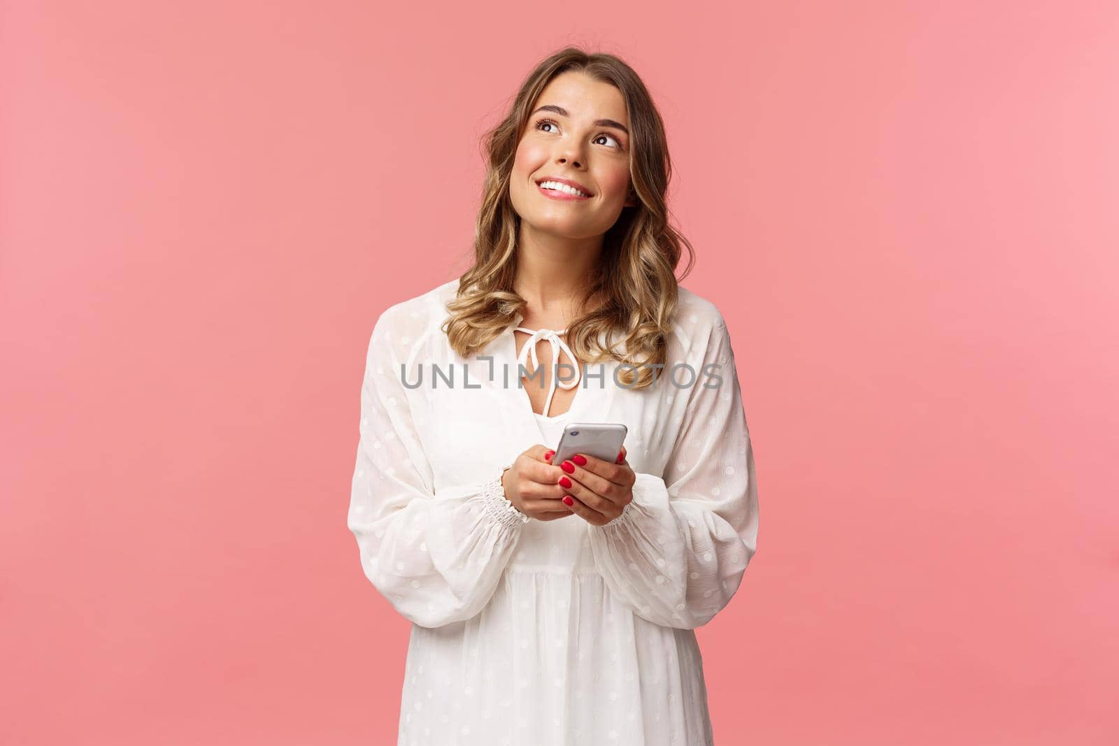 Portrait of dreamy young pretty blond girl fantasizing about something beautiful, looking up thoughtful and happy smiling, holding mobile phone, messaging, shopping online, pink background.