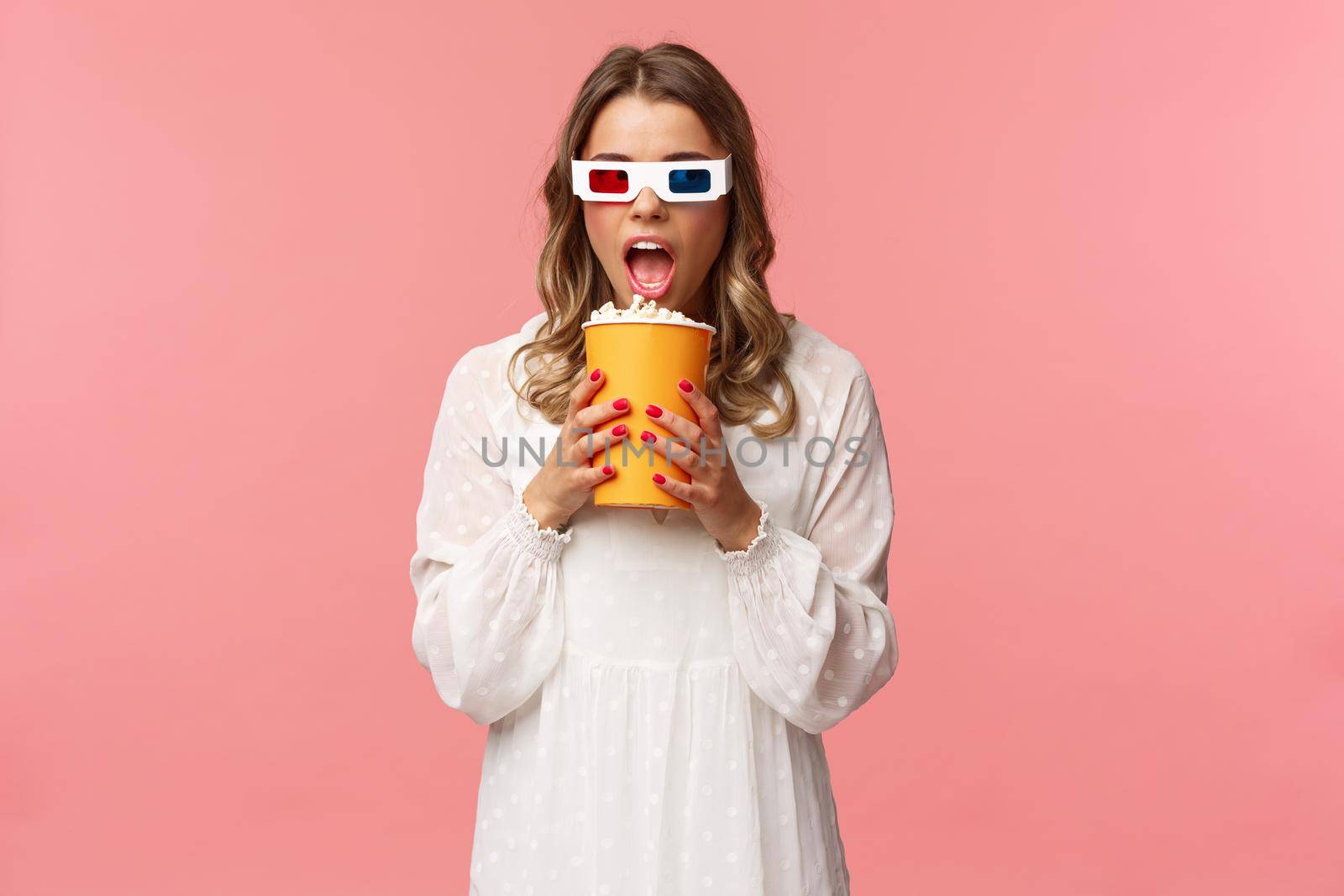 Leisure, going-out and spring concept. Portrait of cute blond funny girl in cinema, eating popcorn from box without hands, looking aside suspicious, wearing 3d glasses, pink background by Benzoix