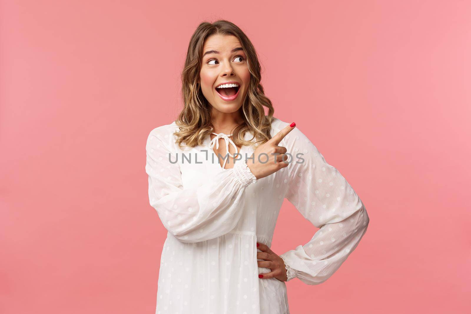 Enthusiastic, happy attractive blond woman in white cute dress, open mouth excited and cheerful looking, pointing upper right corner as best prices discounts ever, stand pink background by Benzoix