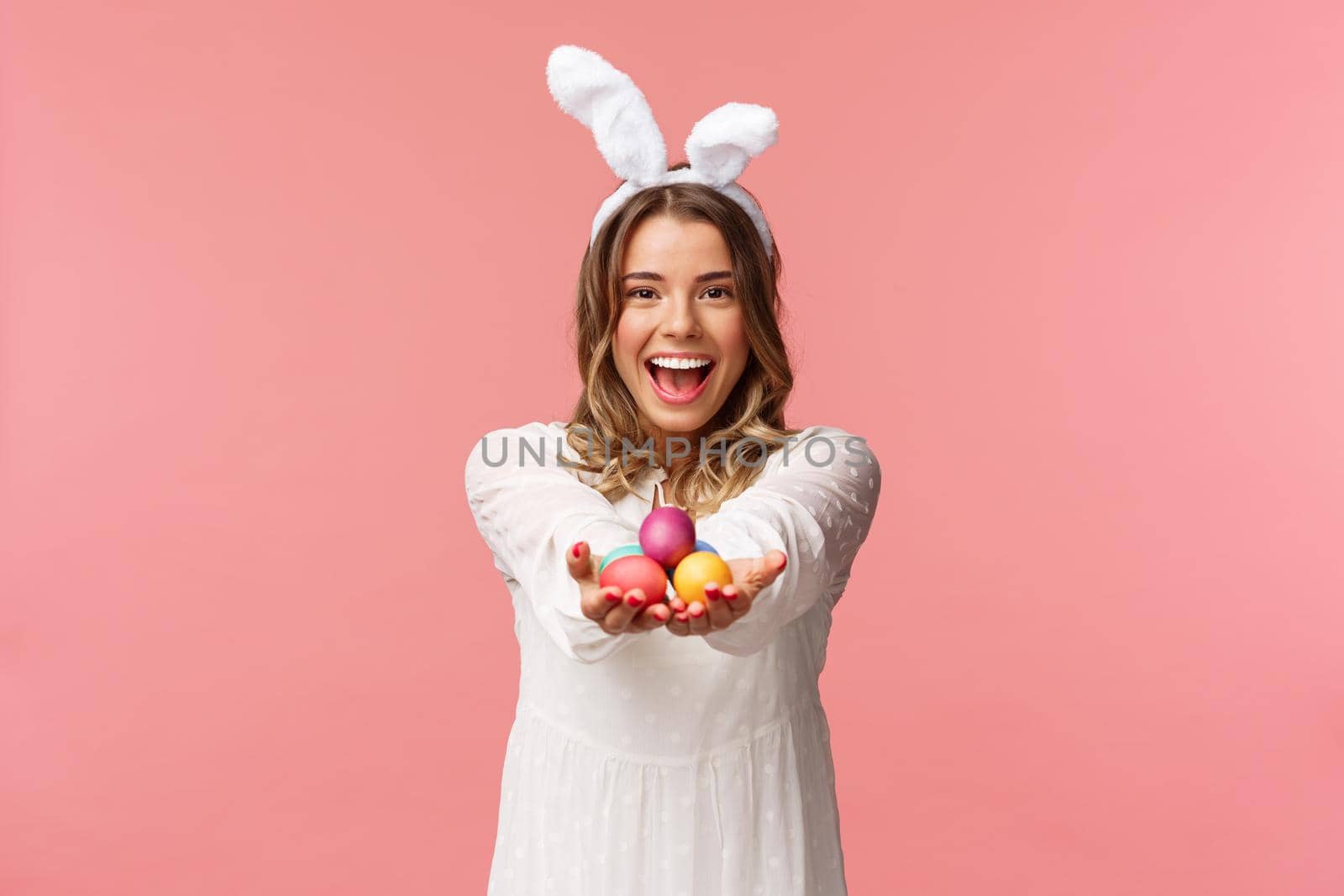 Holidays, spring and party concept. Portrait of lovely, romantic young blond woman in rabbit ears and white dress, giving you painted easter eggs as celebrating orthodox holiday, pink background by Benzoix