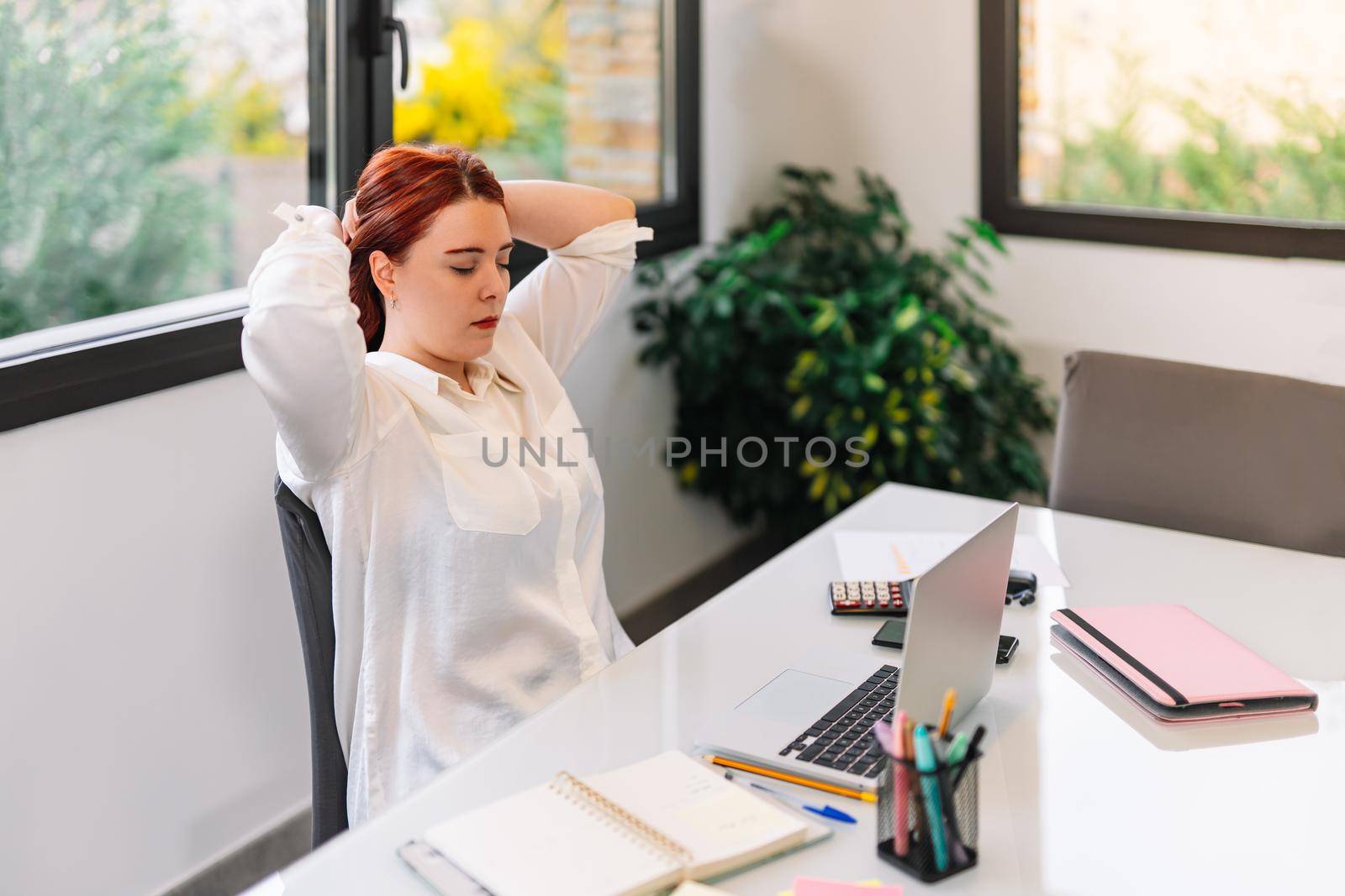 Pretty young woman with red hair massaging her neck while she starts teleworking at home. Self-employed entrepreneur working from home in white shirt. She is working at a light table with a white laptop, calculator, stapler, diaries, notebooks, pens and other devices. Large window in the background with natural light.