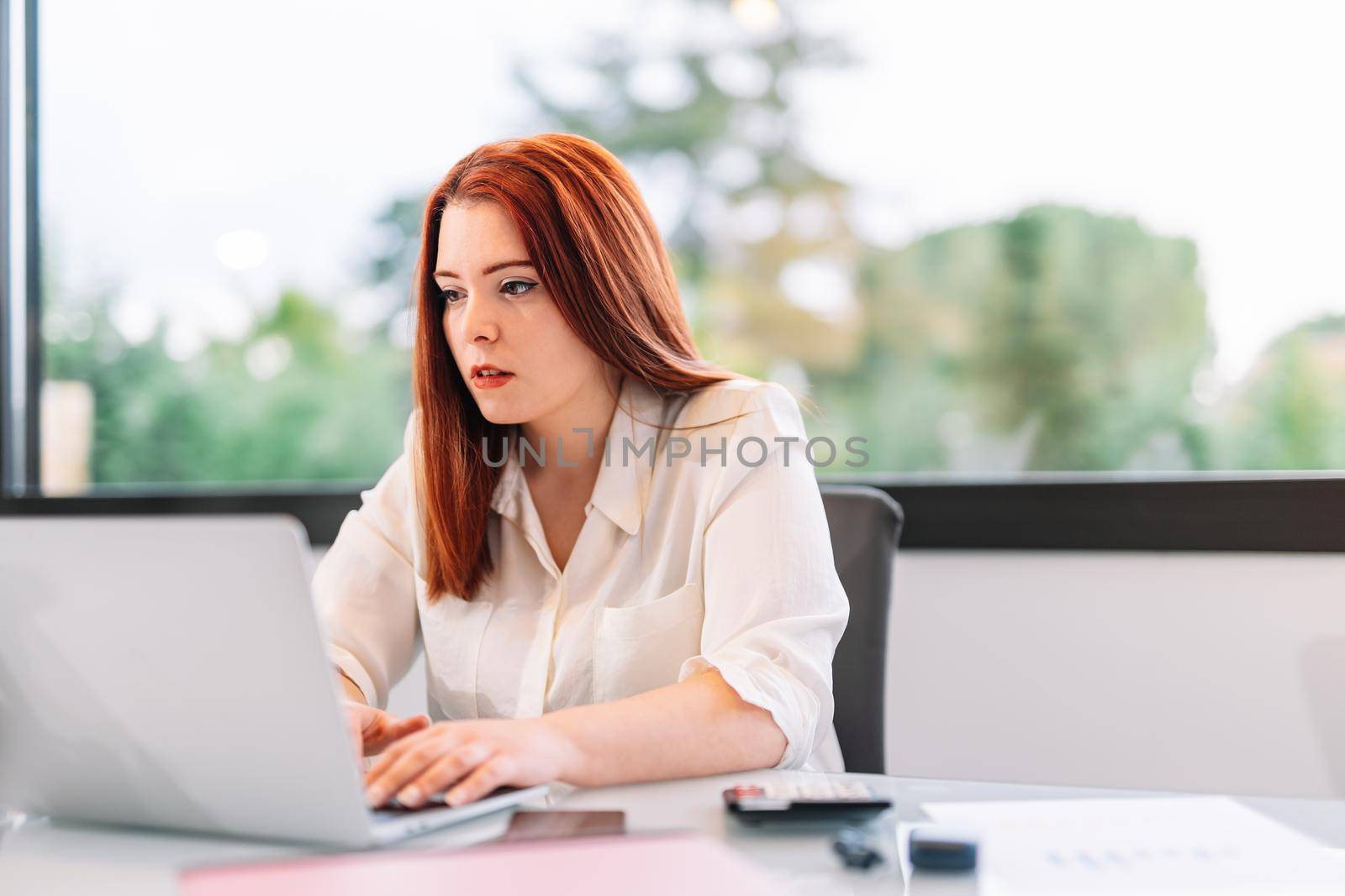 Pretty young red-haired woman in white shirt teleworking on a white laptop from home. She is typing on the laptop while working on her own business. Young entrepreneur and self-employed. She is in an office or at home with a large window behind her with natural light.