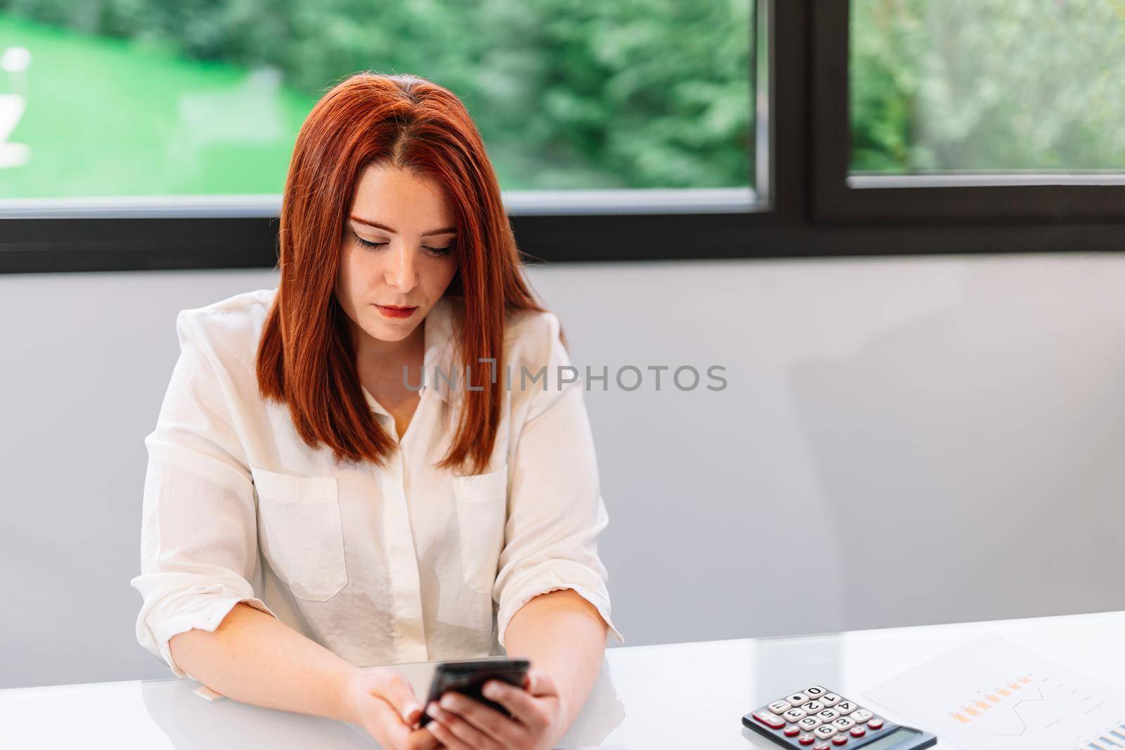 Pretty young red-haired woman looking at her smartphone while teleworking at home. Self-employed entrepreneur and businesswoman working from home or in an office while using her mobile. Large window in background with natural light.