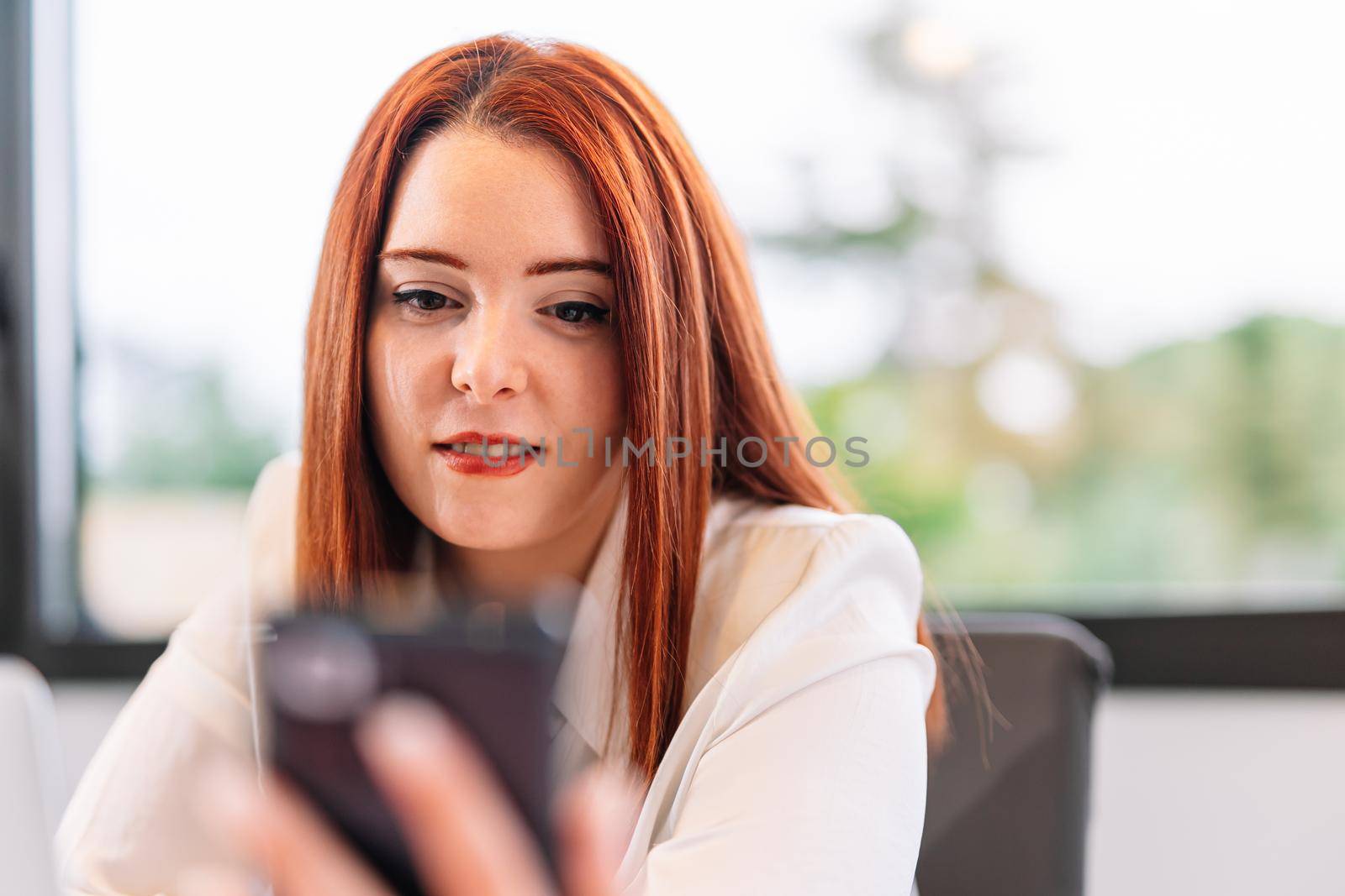 Pretty young red-haired woman looking at her smartphone while teleworking at home. Self-employed entrepreneur and businesswoman working from home or in an office while using her mobile. Large window in background with natural light.