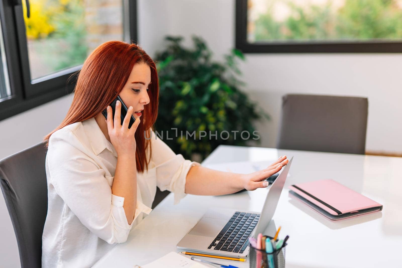 Pretty young beautiful woman with red hair and white shirt teleworking from home talking on her smartphone while using her laptop. She is making a phone call while looking at the laptop screen. Young self-employed businesswoman and entrepreneur working from home with large window in background and natural light.