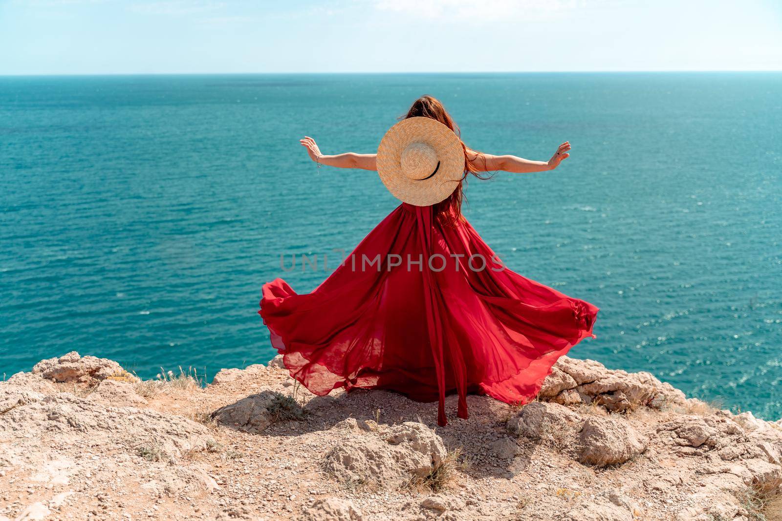 A woman, rear view in a red flying dress fluttering in the wind, a girl in a fluttering dress on the background of the sea. A straw hat hangs at the back of the neck