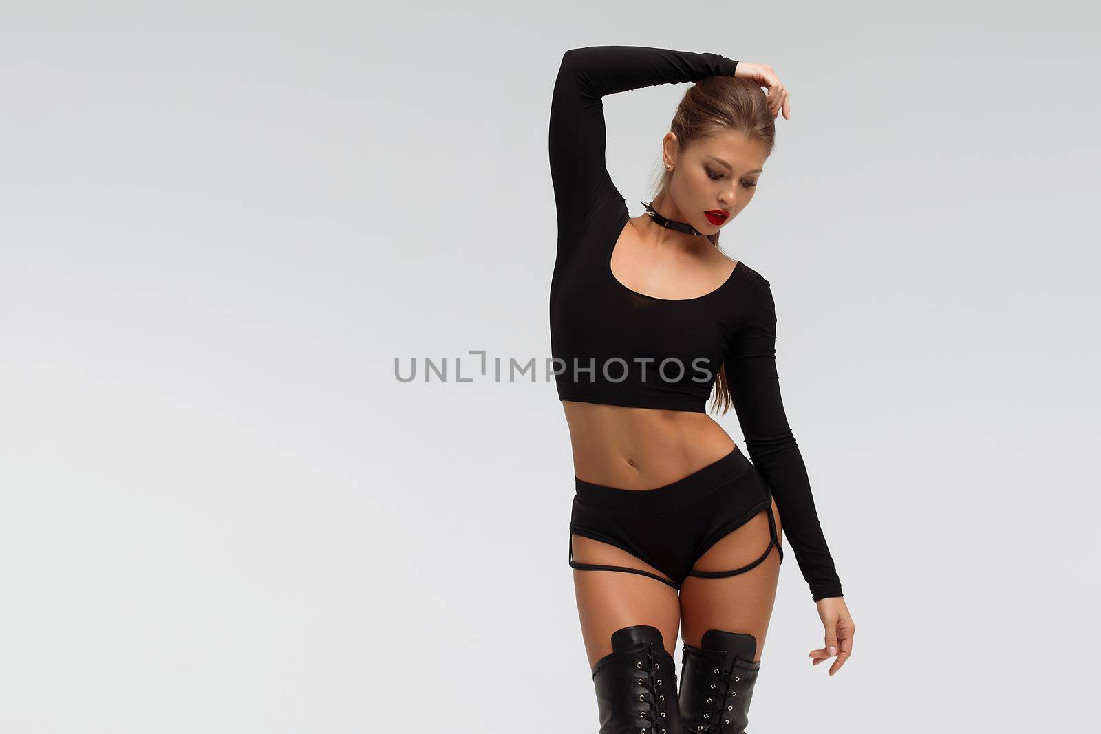 Full body of provocative young slim female with red lips wearing black underwear and high heeled boots standing against white background