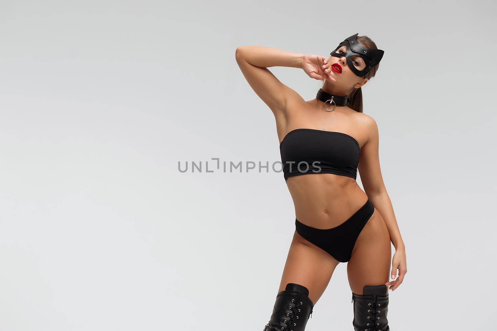 Slim tanned young female in black underwear and mask with collar on neck touching head while standing against white background