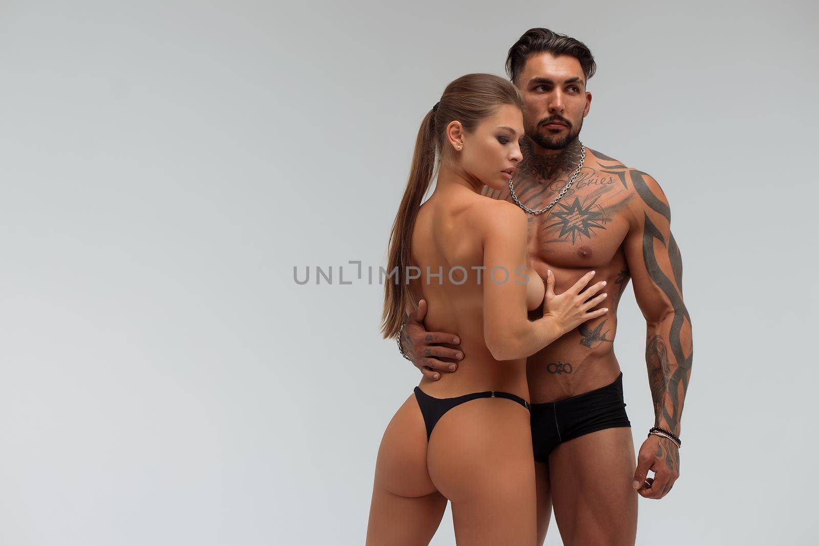 Muscular tattooed man embracing slim topless woman in panties on gray background