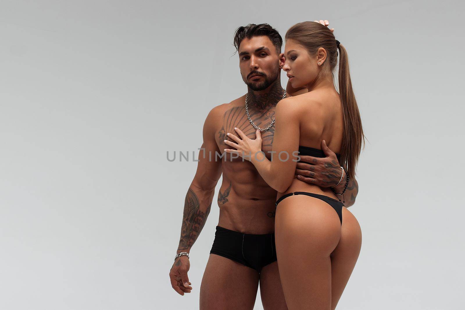 Muscular tattooed man embracing slim topless woman in panties on gray background