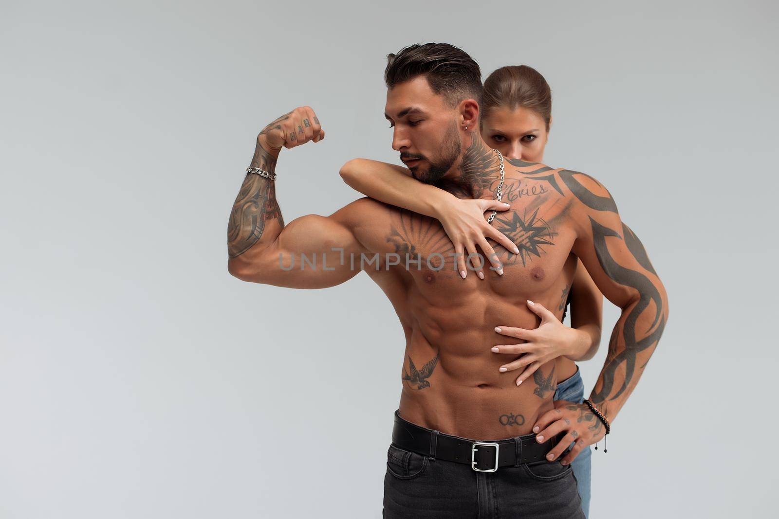 Strong shirtless male embracing and covering breast of attractive topless female on gray background