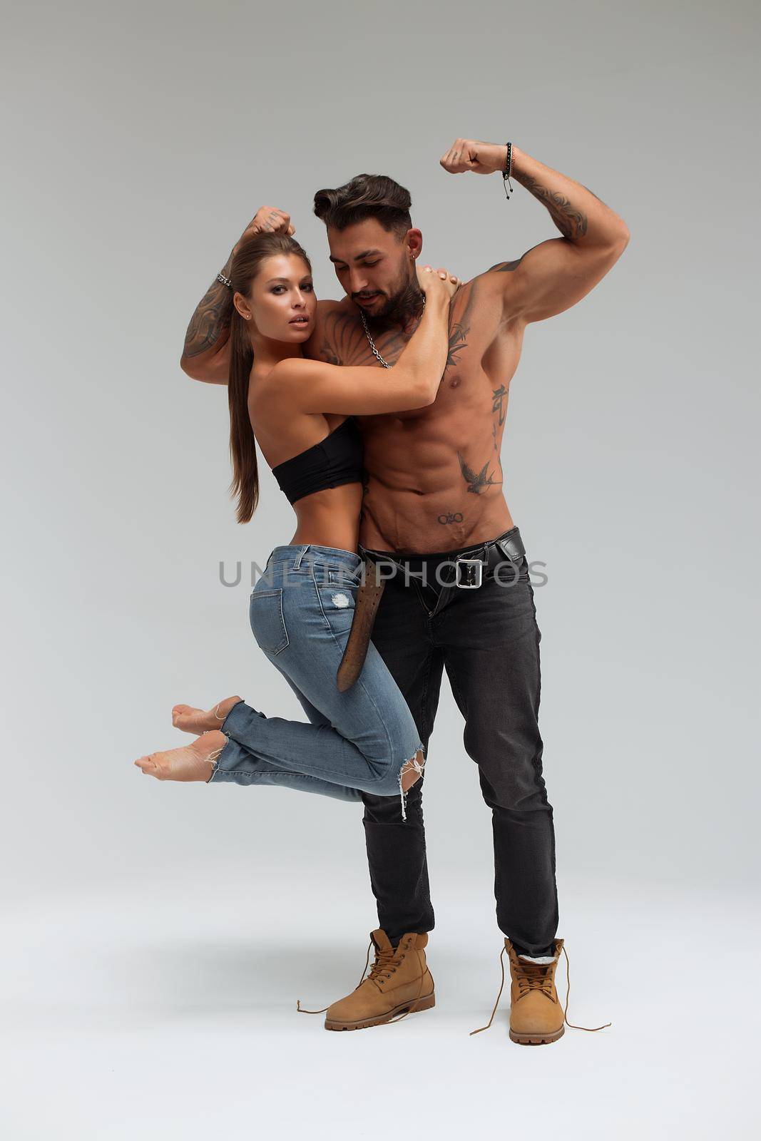 Attractive female embracing handsome shirtless male with tattoos from behind and looking at camera against gray background