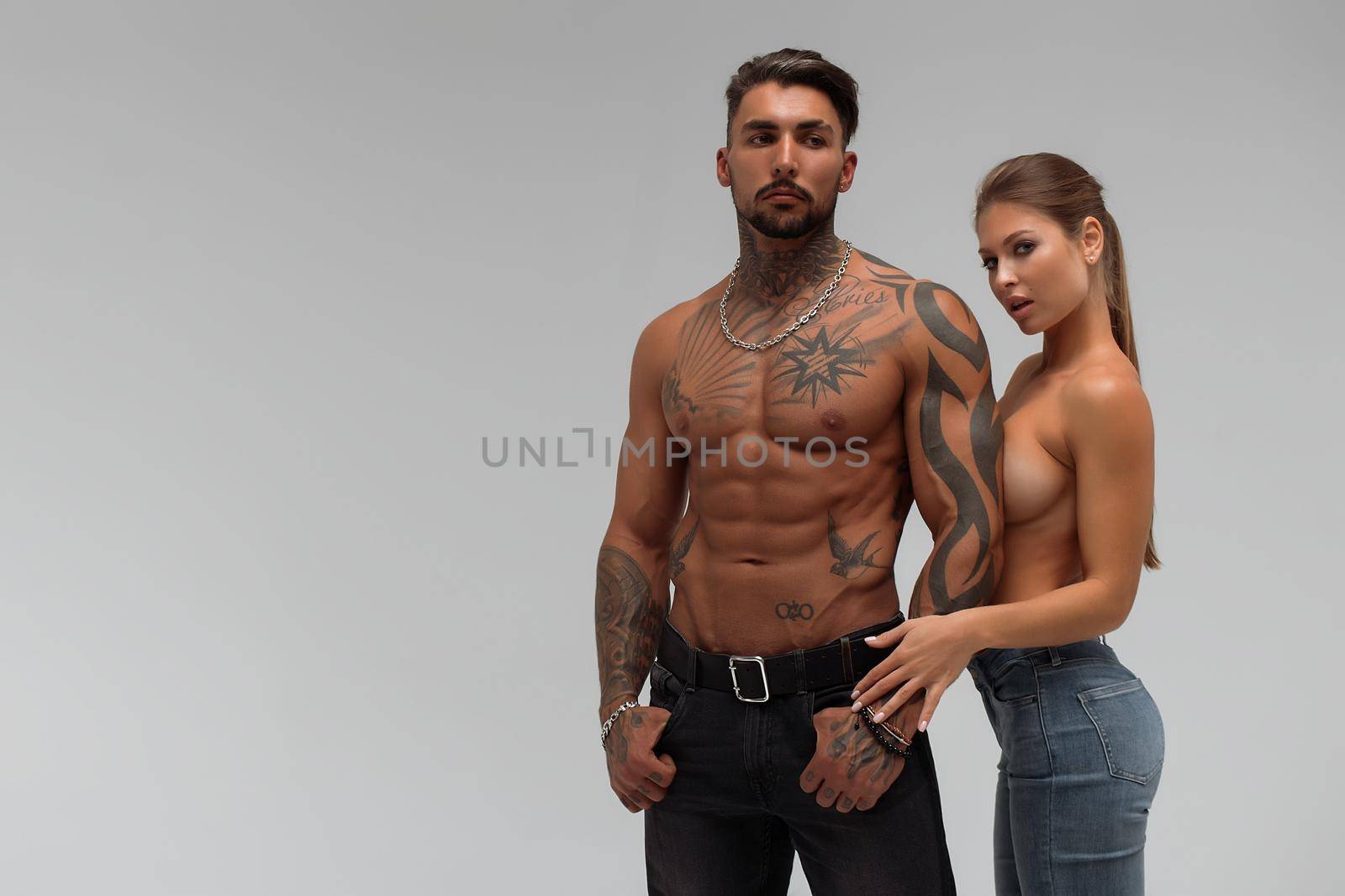 Muscular tattooed man embracing slim topless woman in panties on gray background