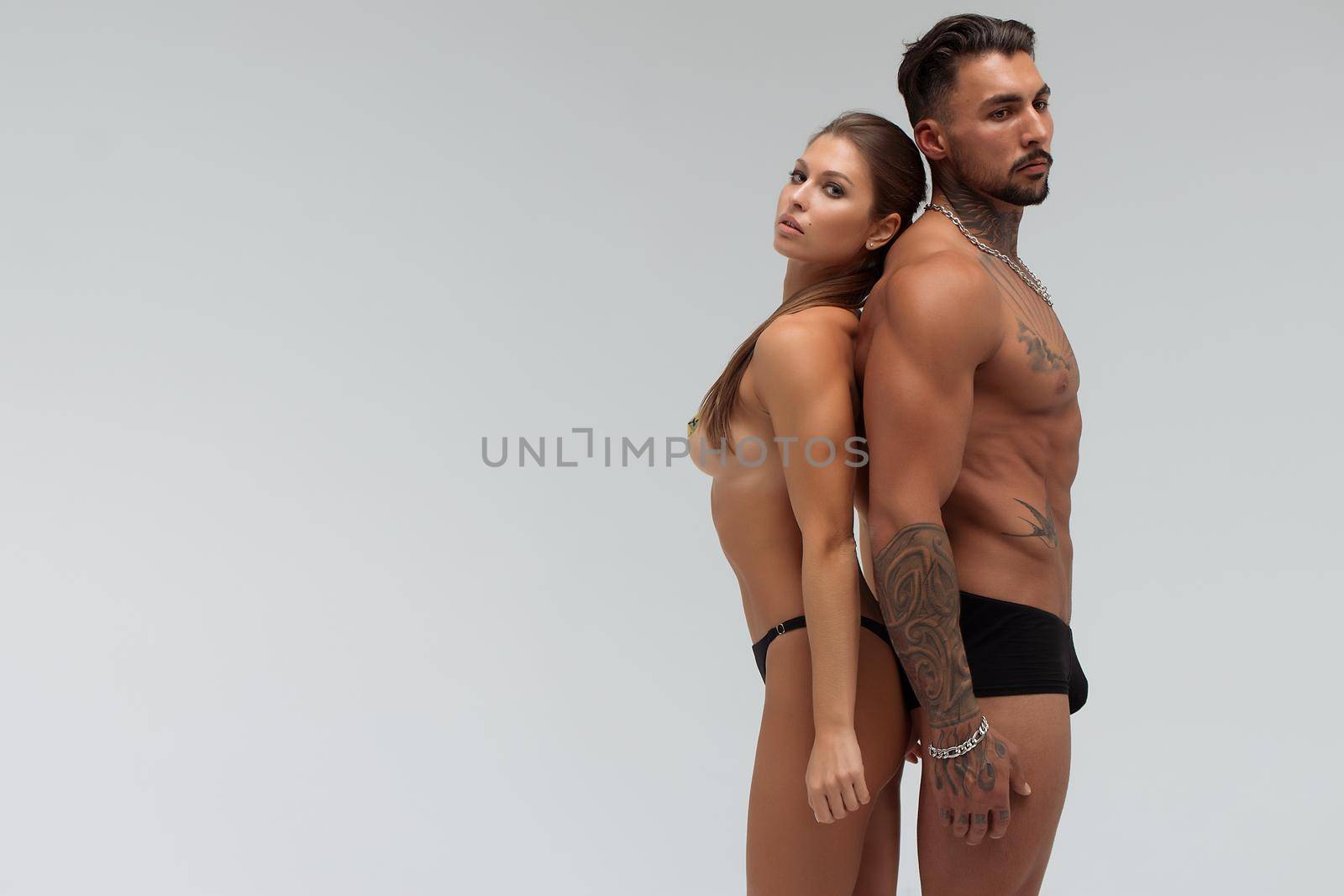 Strong shirtless male embracing and covering breast of attractive topless female on gray background