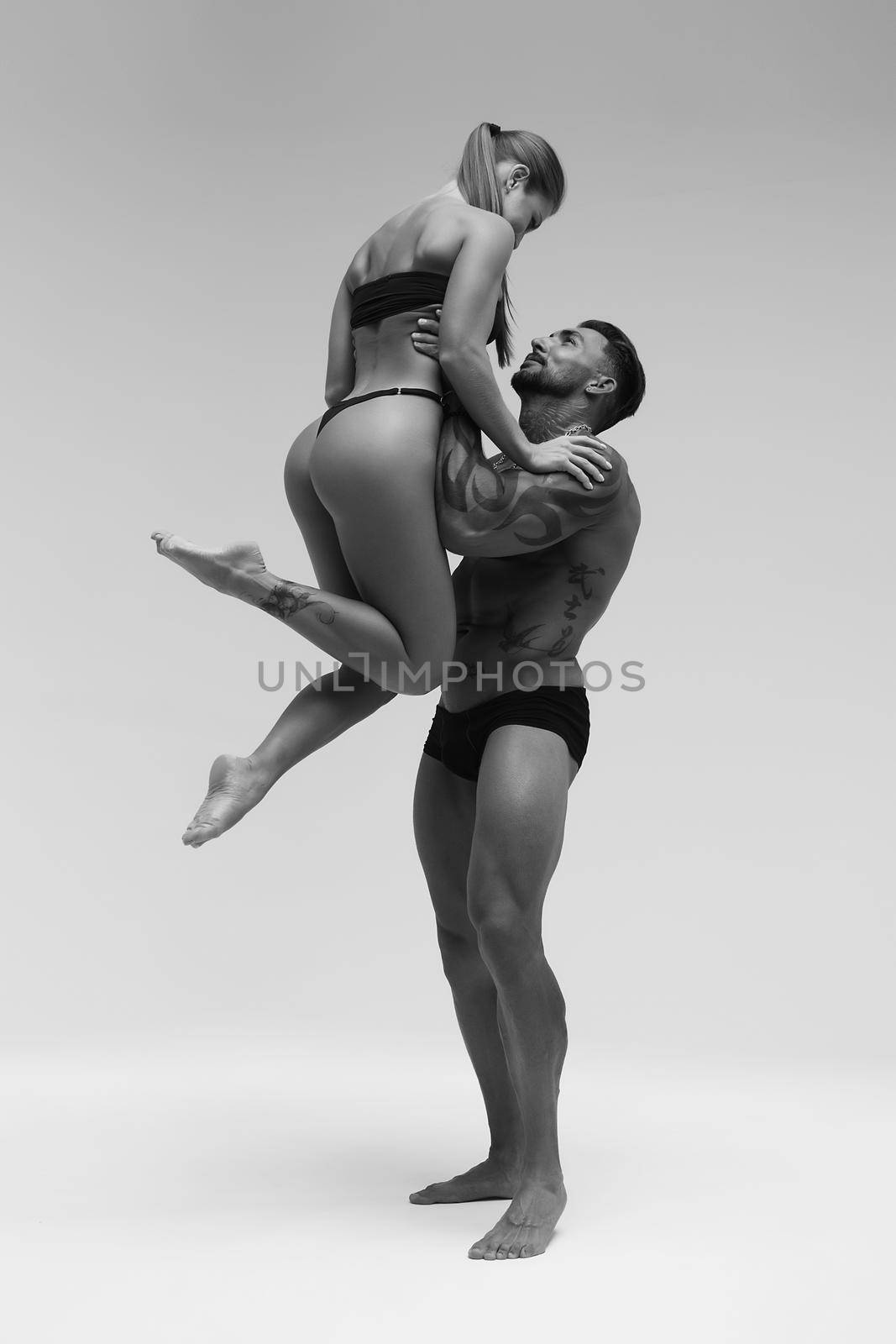 Black and white side view of topless woman and shirtless man looking at camera against gray background