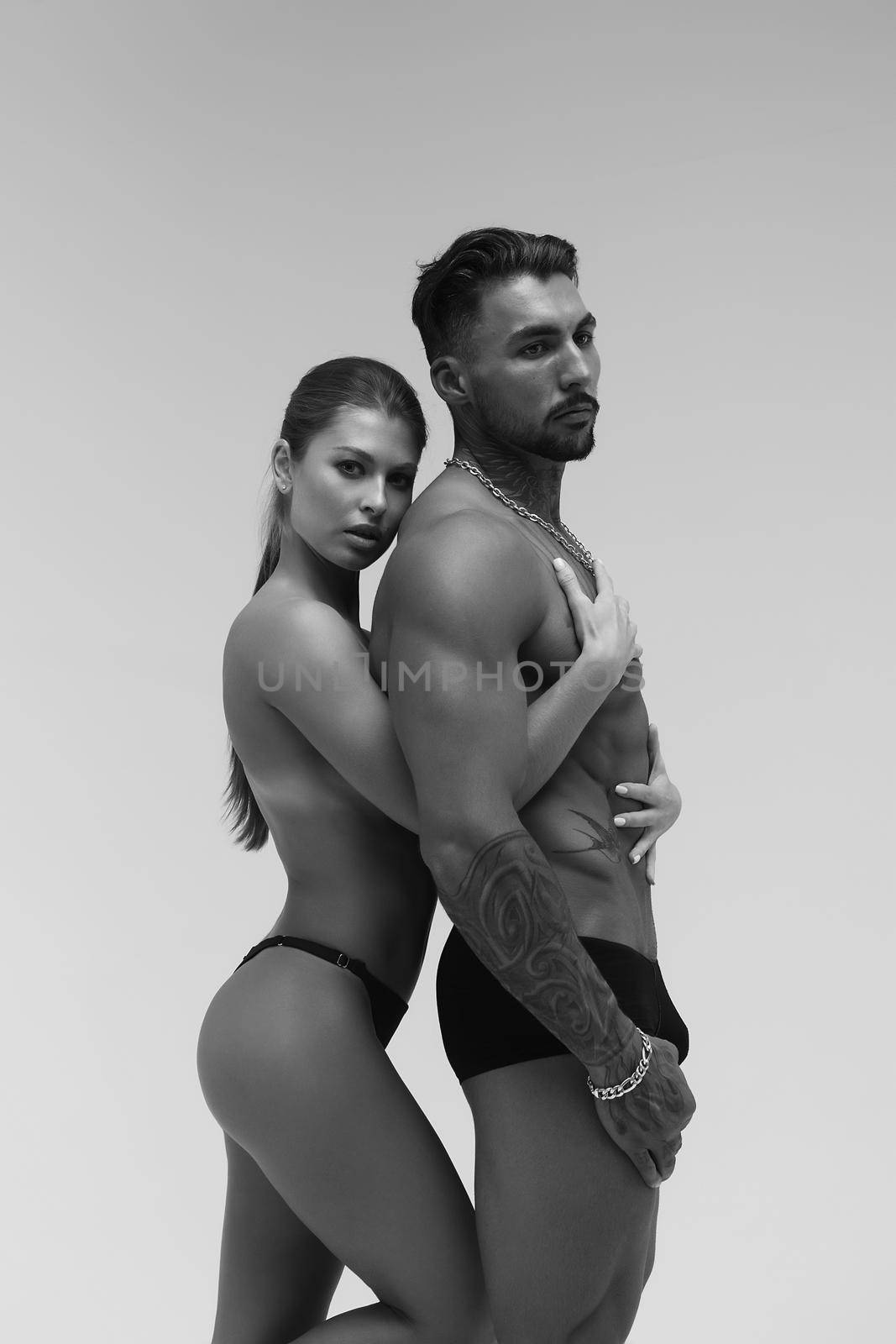 Black and white side view of topless woman and shirtless man looking at camera against gray background