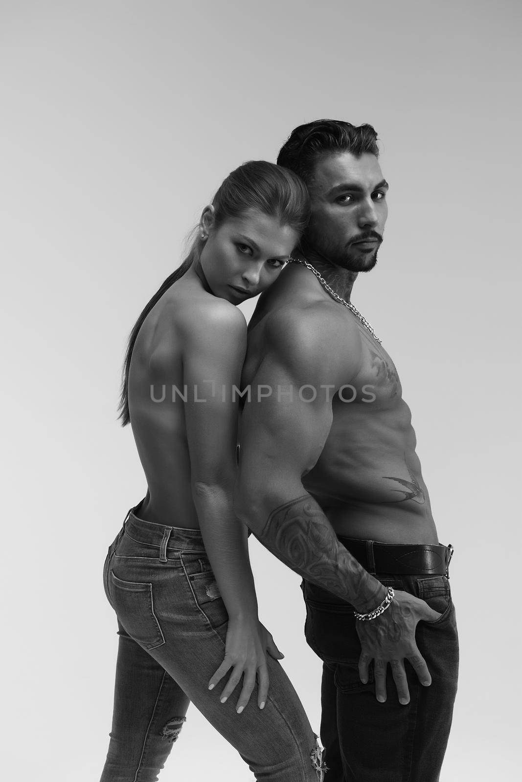 Black and white side view of topless woman and shirtless man looking at camera against gray background