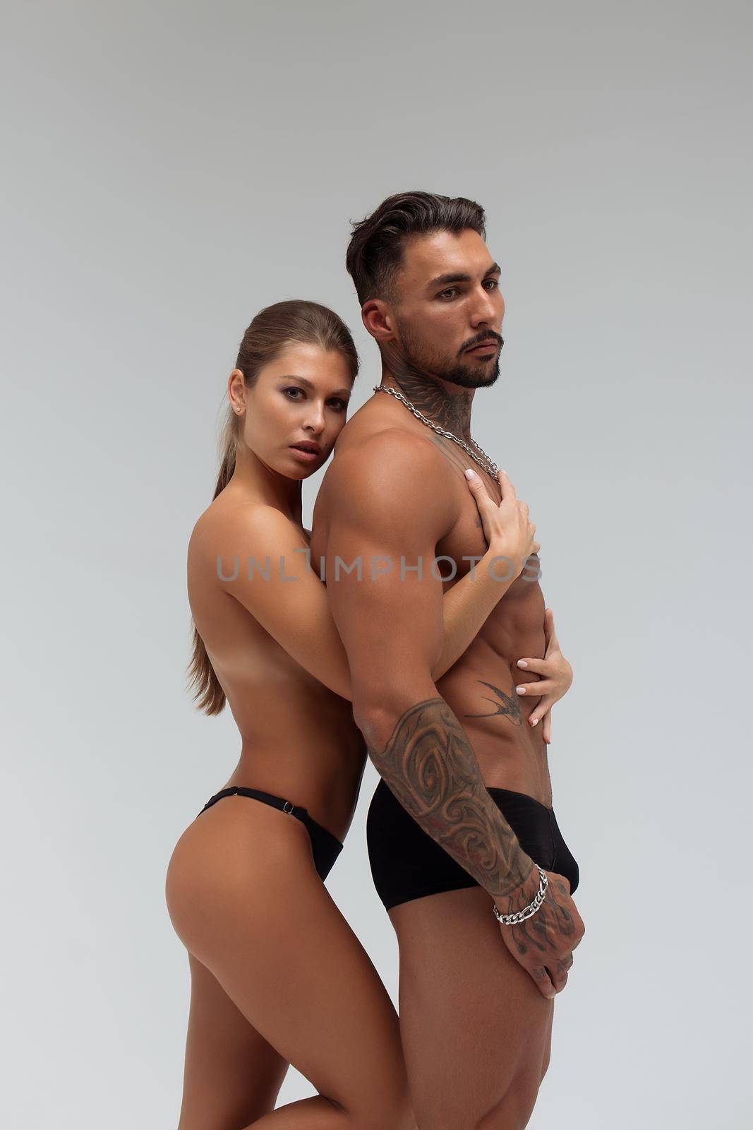 Attractive female embracing handsome shirtless male with tattoos from behind and looking at camera against gray background