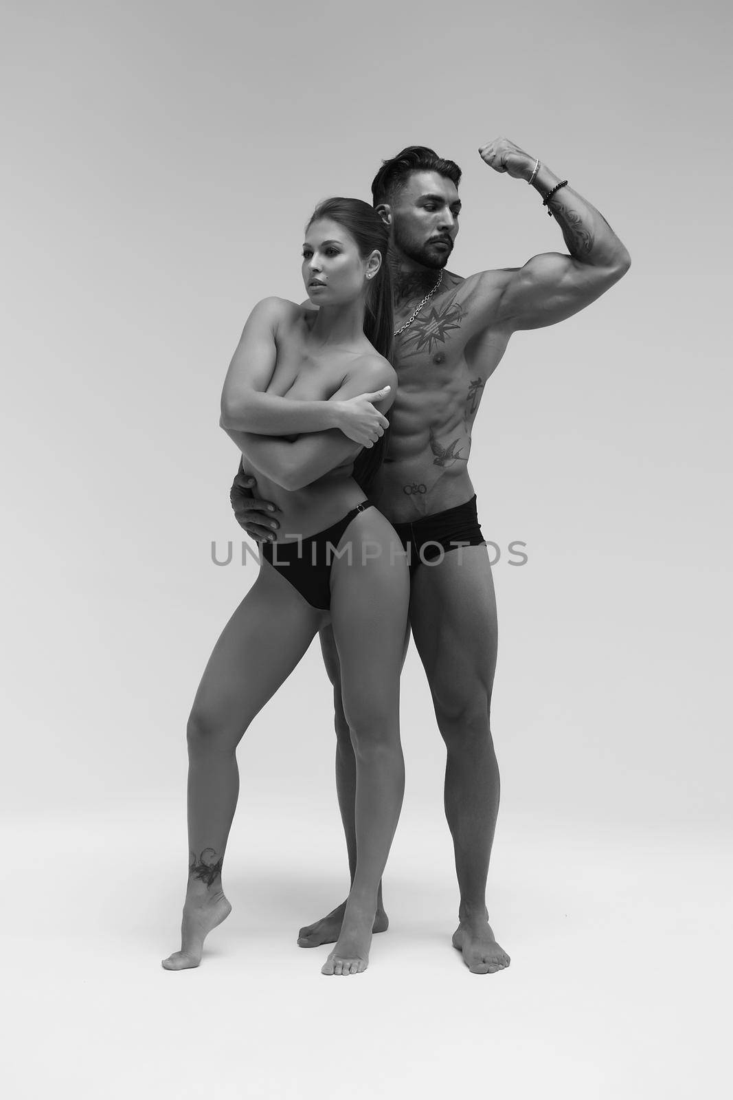 Black and white side view of topless woman and shirtless man looking at camera against gray background