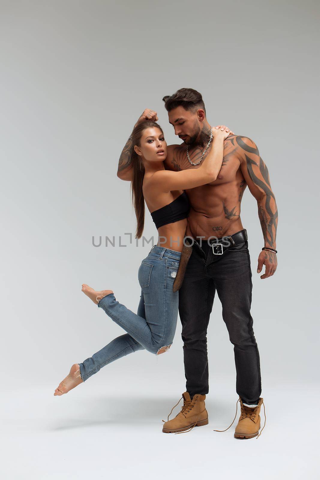 Attractive female embracing handsome shirtless male with tattoos from behind and looking at camera against gray background