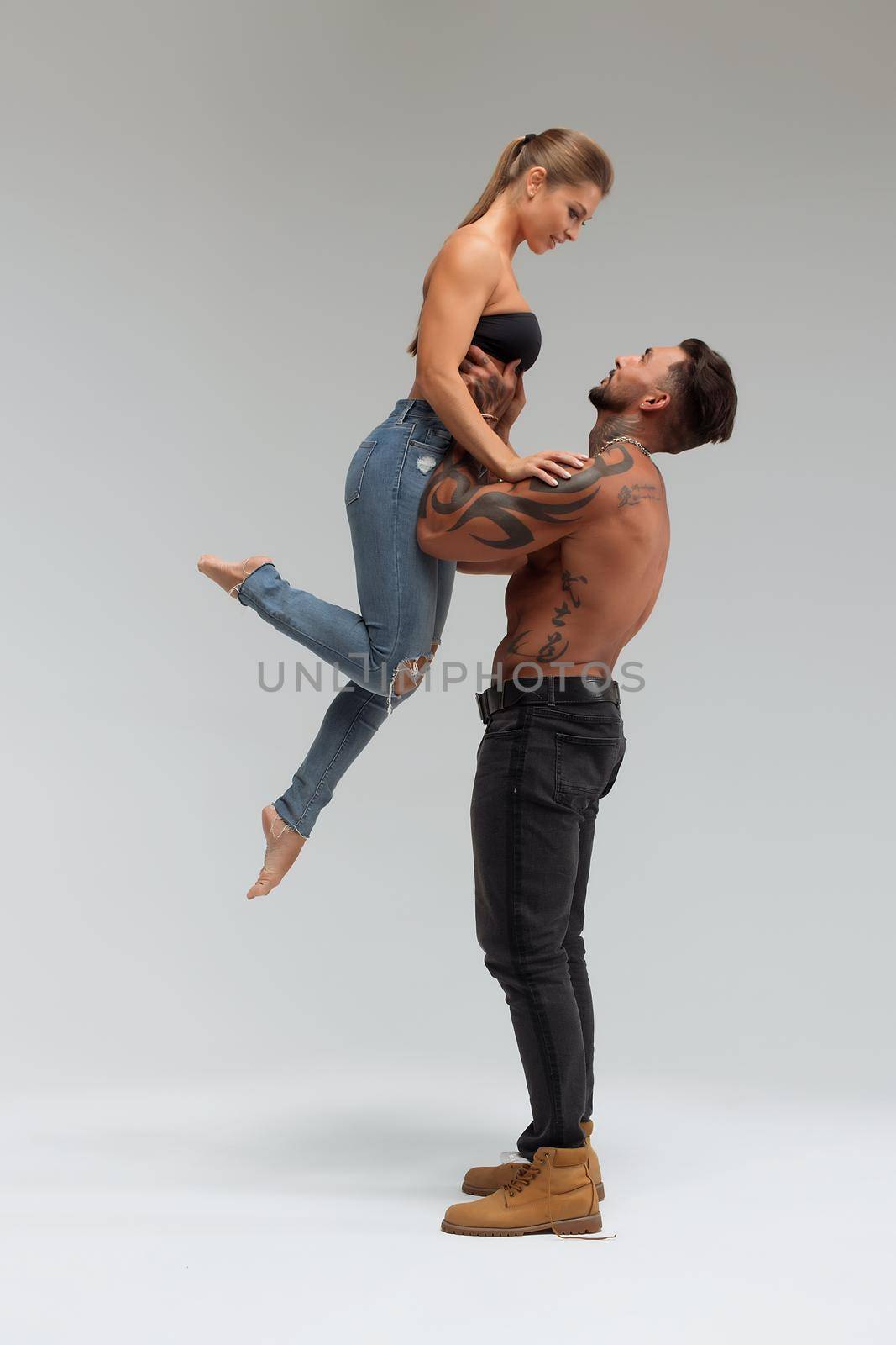 Attractive female embracing handsome shirtless male with tattoos from behind and looking at camera against gray background