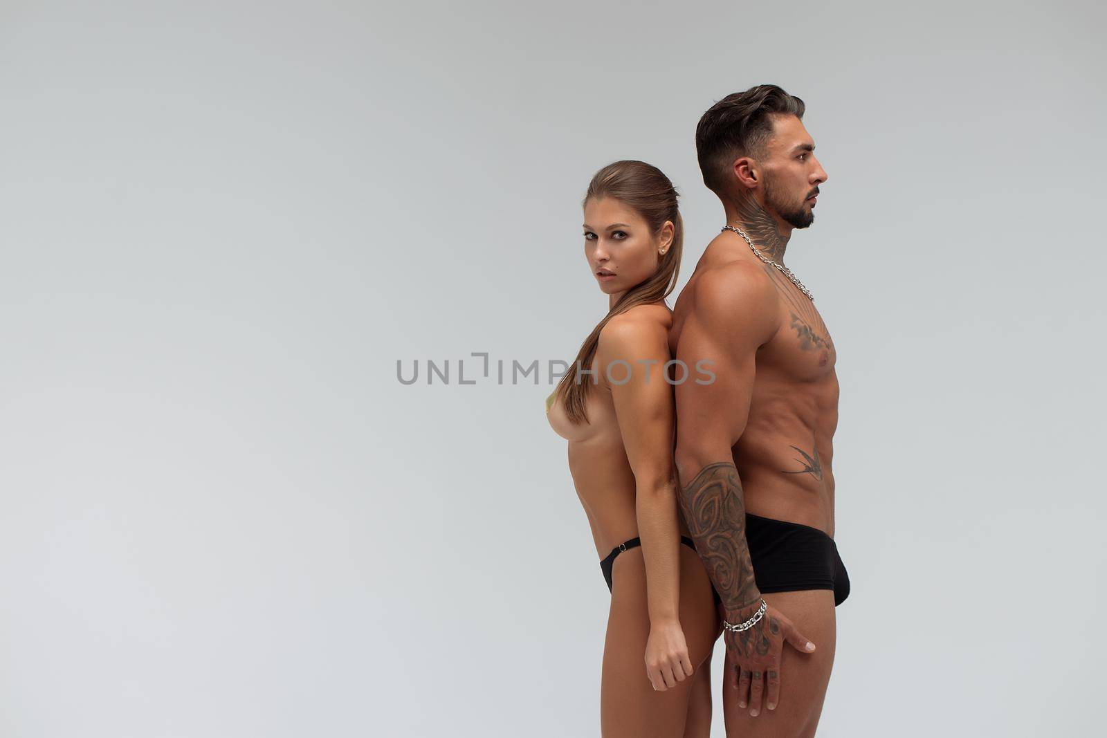 Muscular tattooed man embracing slim topless woman in panties on gray background