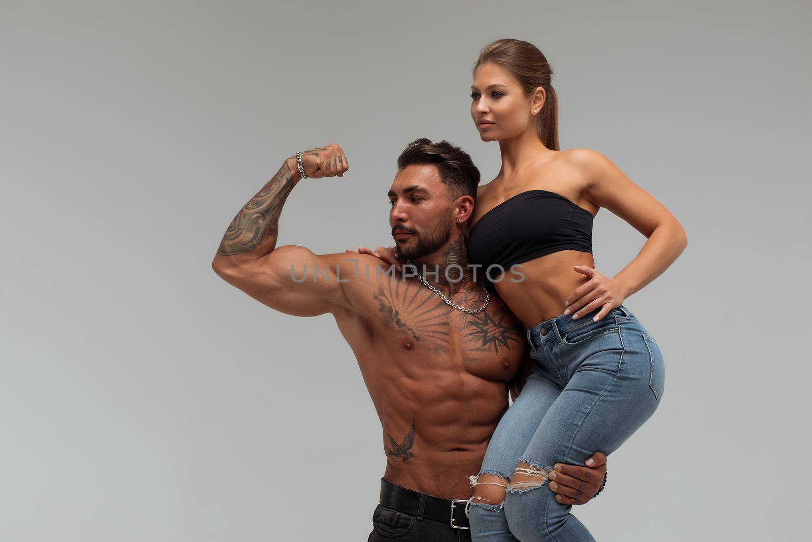 Strong shirtless male embracing and covering breast of attractive topless female on gray background