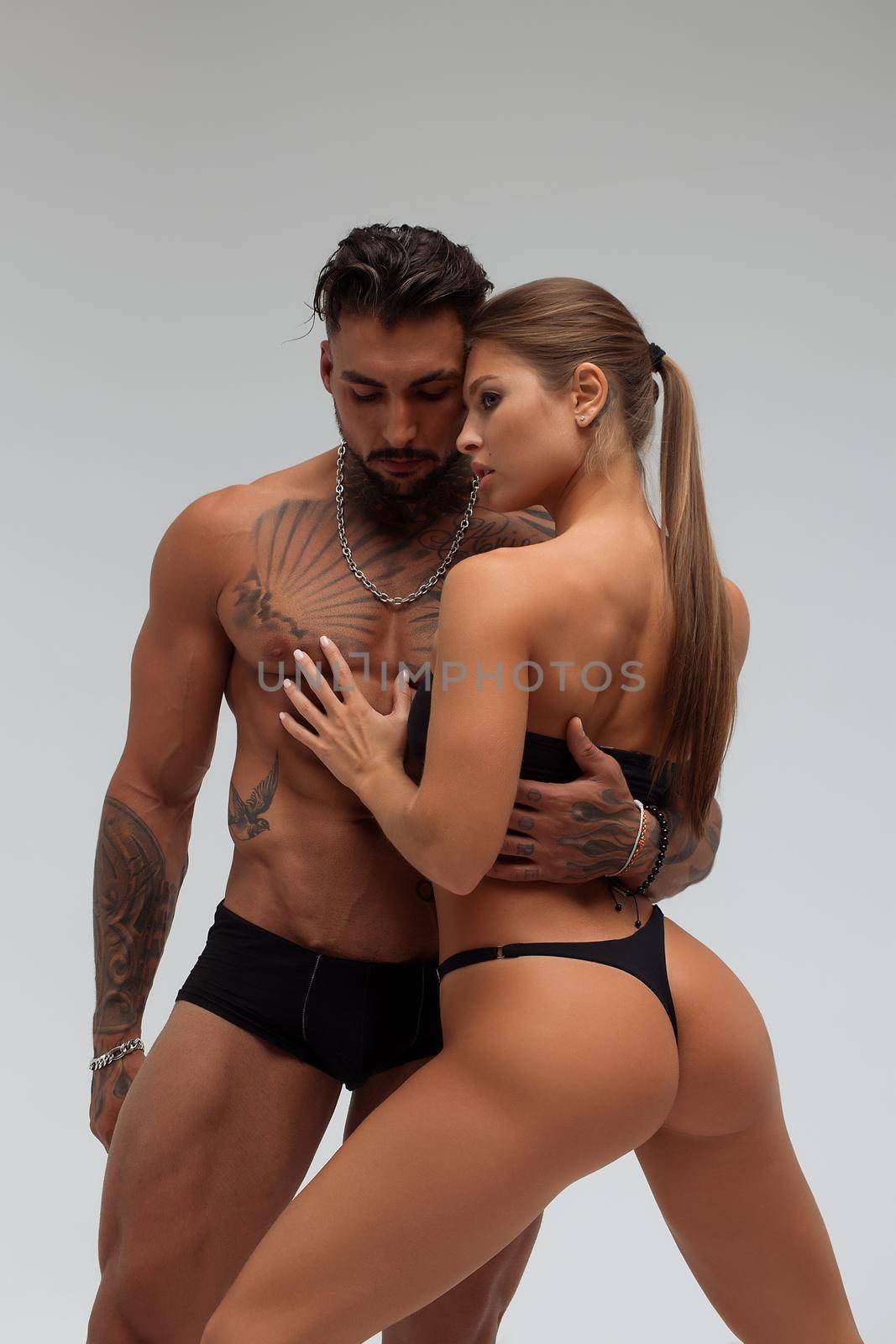 Attractive female embracing handsome shirtless male with tattoos from behind and looking at camera against gray background