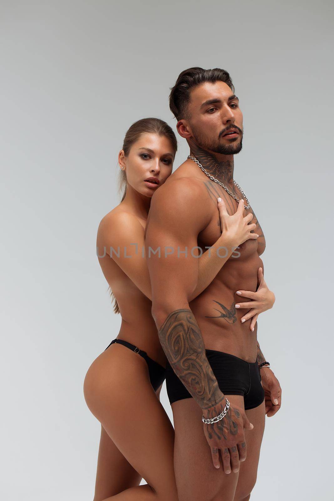 Attractive female embracing handsome shirtless male with tattoos from behind and looking at camera against gray background