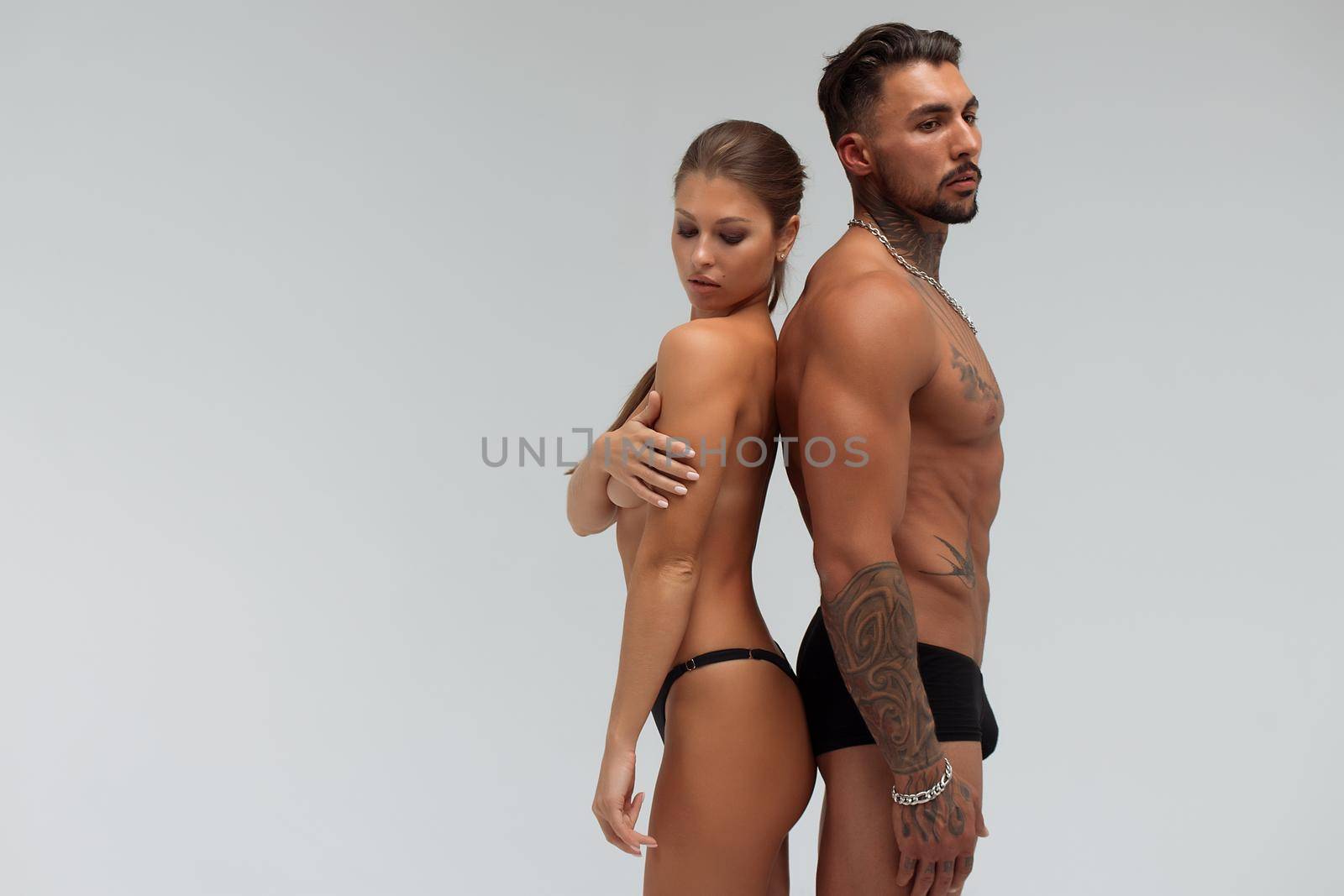 Muscular tattooed man embracing slim topless woman in panties on gray background