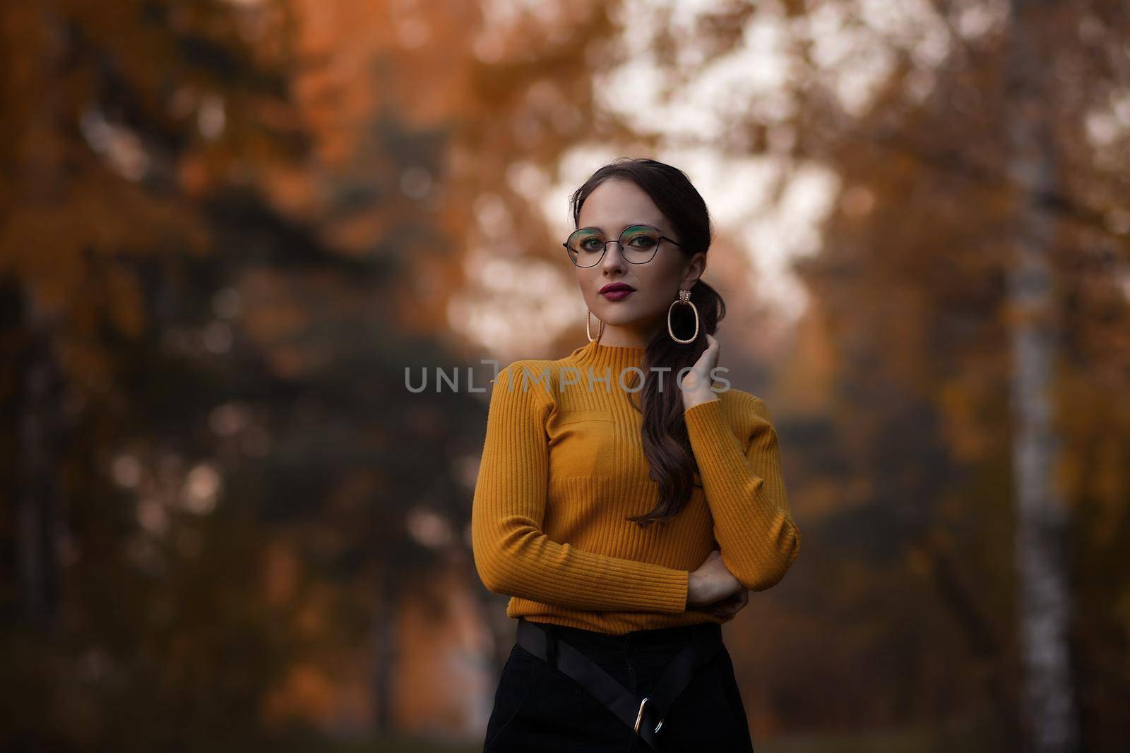 Young long haired brunette in yellow knitted jumper and eyeglasses looking at camera while standing against blurred foliage of autumn trees in park