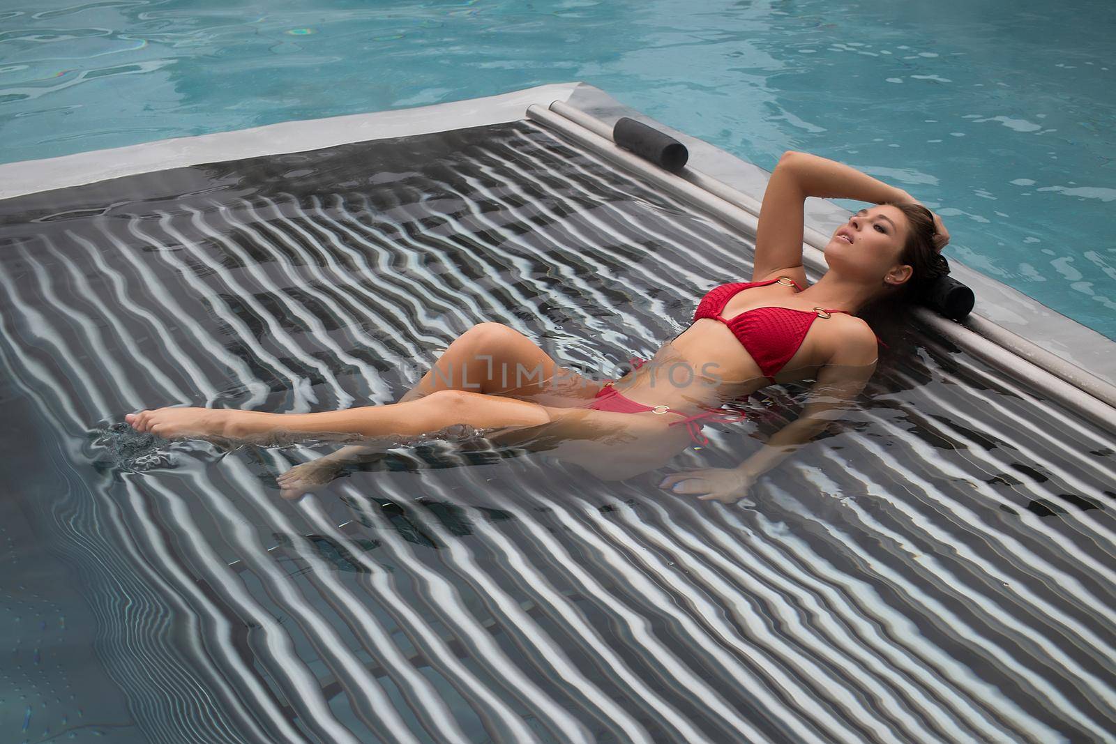 High angle of slim barefoot woman in red swimwear lying on grates in clean water of pool on resort