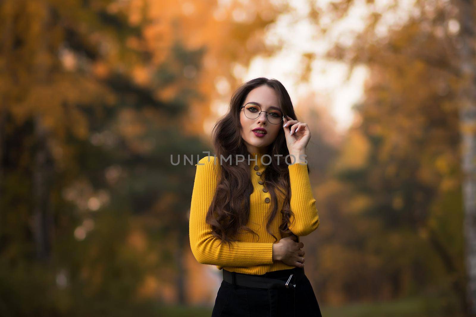 Young long haired brunette in yellow knitted jumper and eyeglasses looking at camera while standing against blurred foliage of autumn trees in park