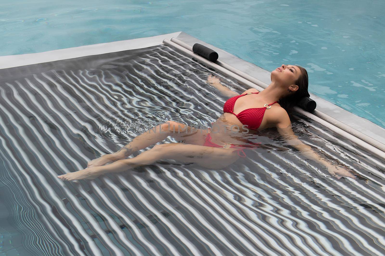 High angle of slim barefoot woman in red swimwear lying on grates in clean water of pool on resort