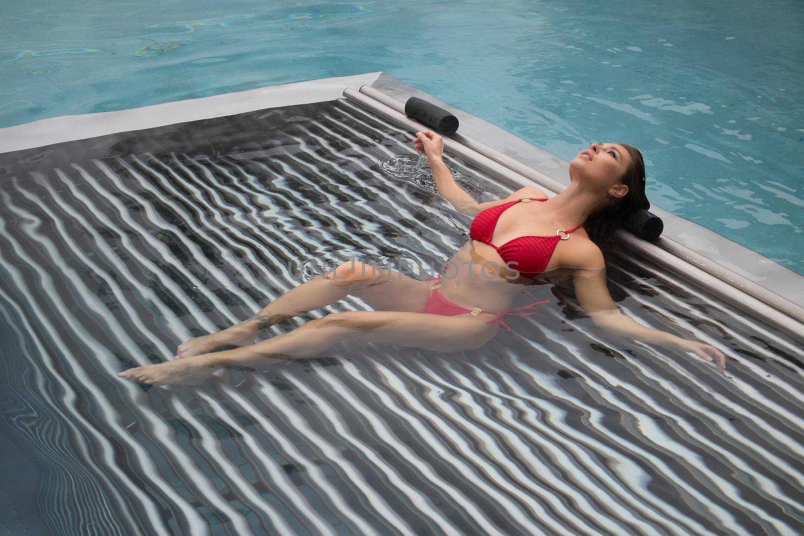 High angle of slim barefoot woman in red swimwear lying on grates in clean water of pool on resort