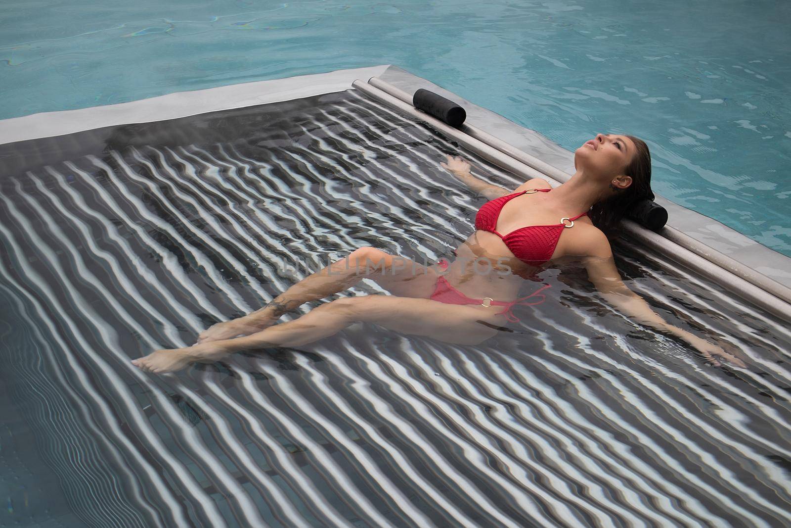 High angle of slim barefoot woman in red swimwear lying on grates in clean water of pool on resort