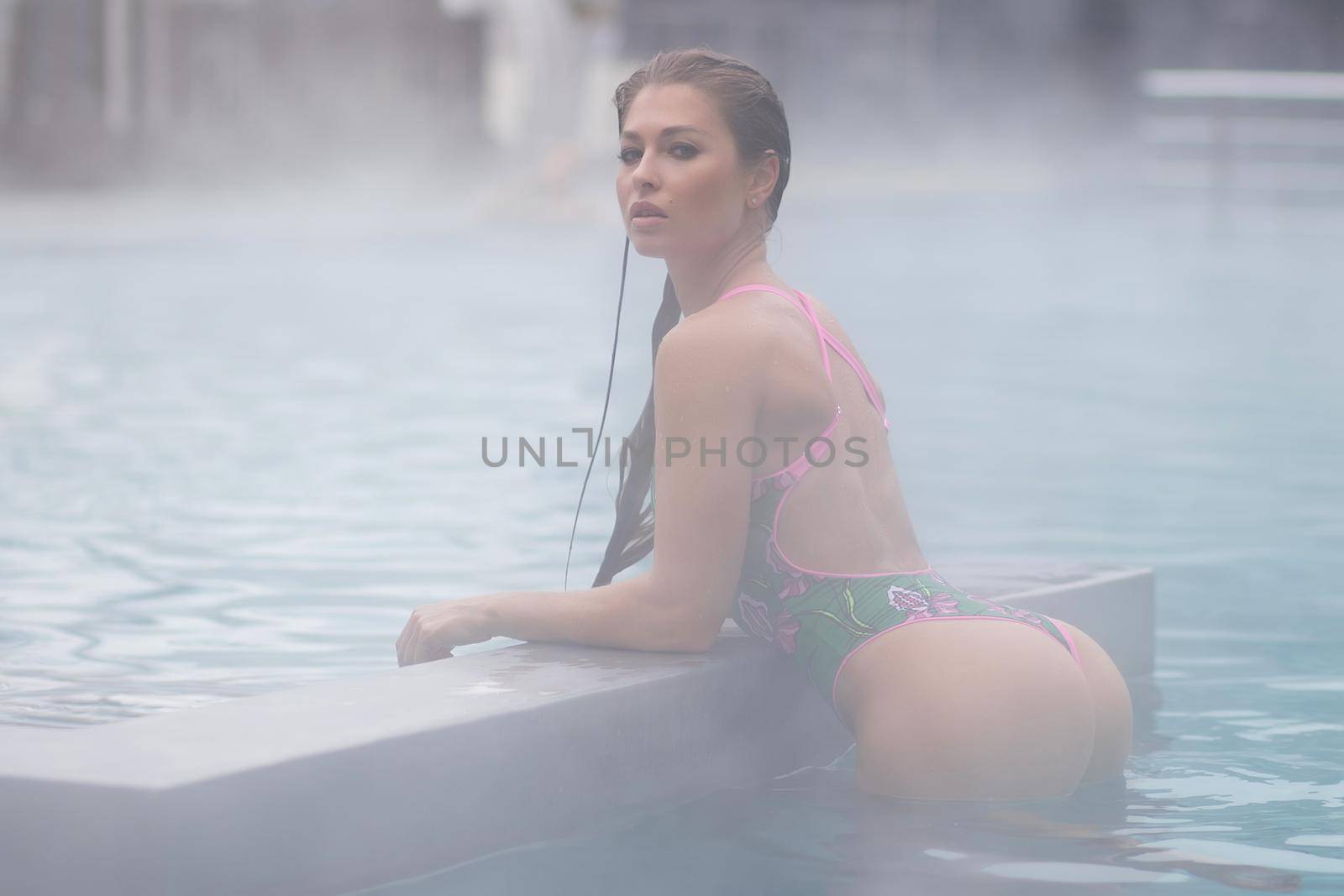 Side view of sensual slim female in green bikini leaning on border and looking at camera in warm water of hot spring