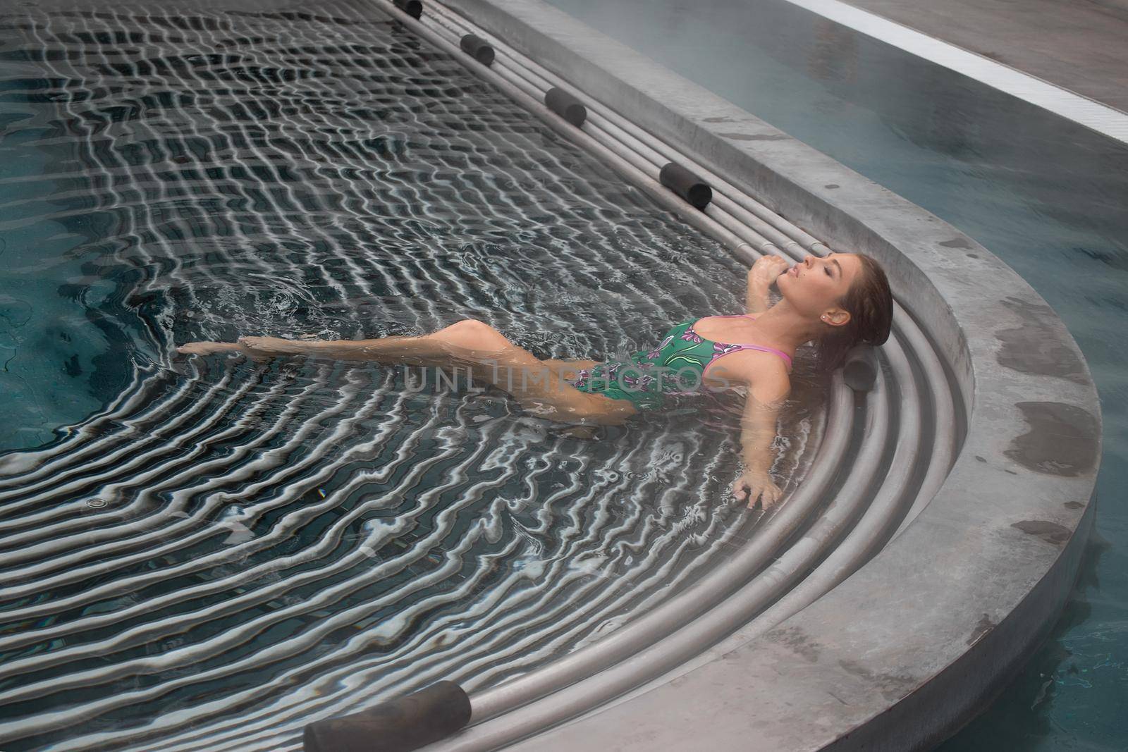 High angle of slim barefoot woman in green swimwear lying on grates in clean water of pool on resort