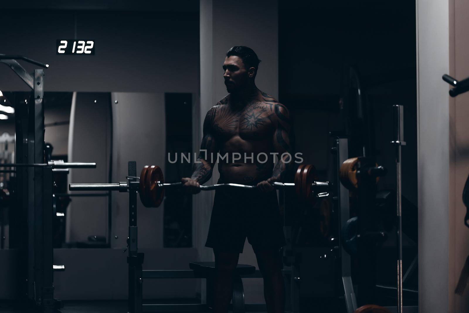 Muscular male athlete with tattooed naked torso standing in dark gym and doing dumbbell curls during workout